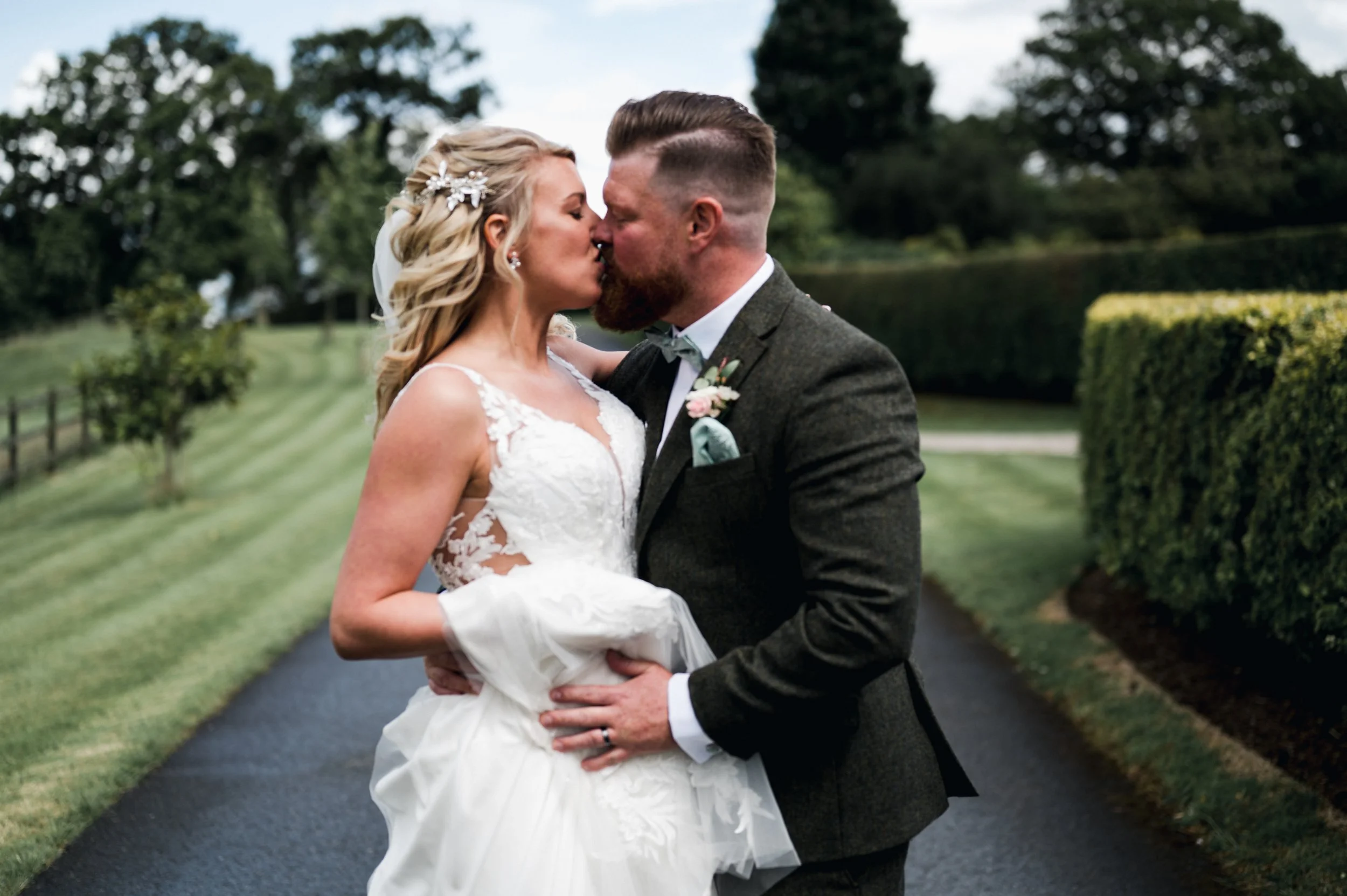 A newlywed couple sharing a kiss outdoors on a cloudy day, with the bride in a white lace wedding gown and the groom in a grey suit with a boutonniere grange barn Whitchurch, Gareth roy photography