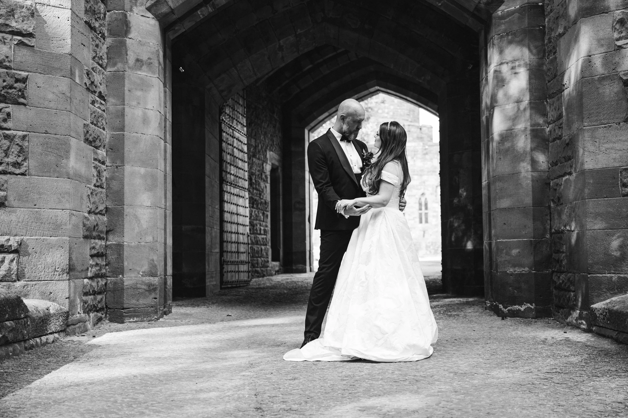 Peckforton Castle - black and white image of bride and groom in wedding holding each others hands in an archway