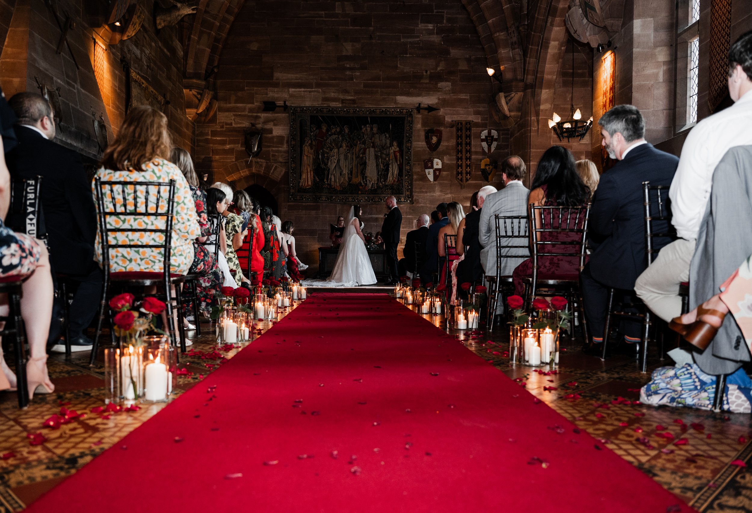 Peckforton Castle - bride and groom in wedding attire at the bottom of the aisle during wedding ceremony with wedding guests sat in the pew
