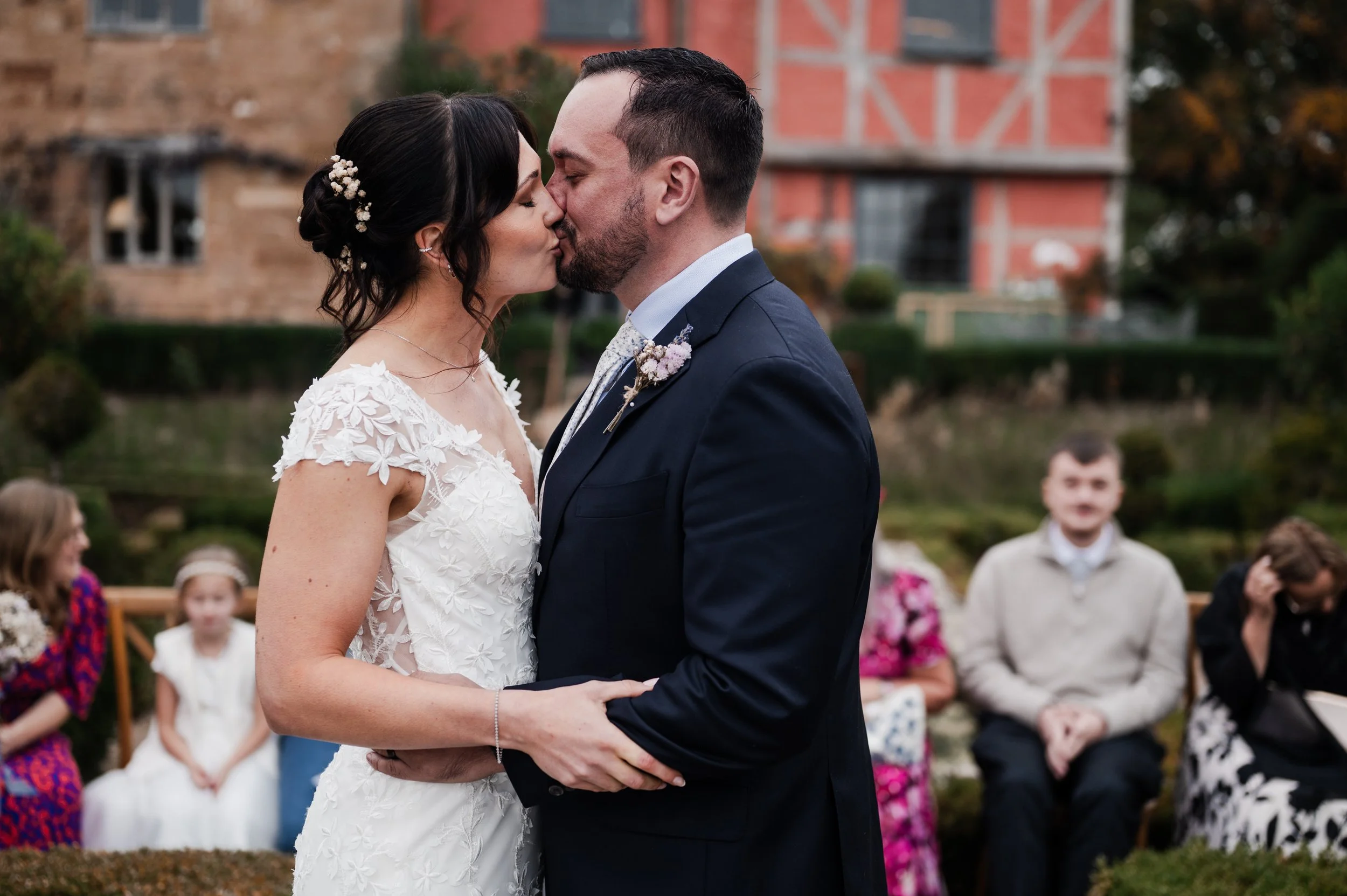 A couple in wedding attire kissing outdoors with wedding guests seated in the background pauntley court, Gareth roy photography