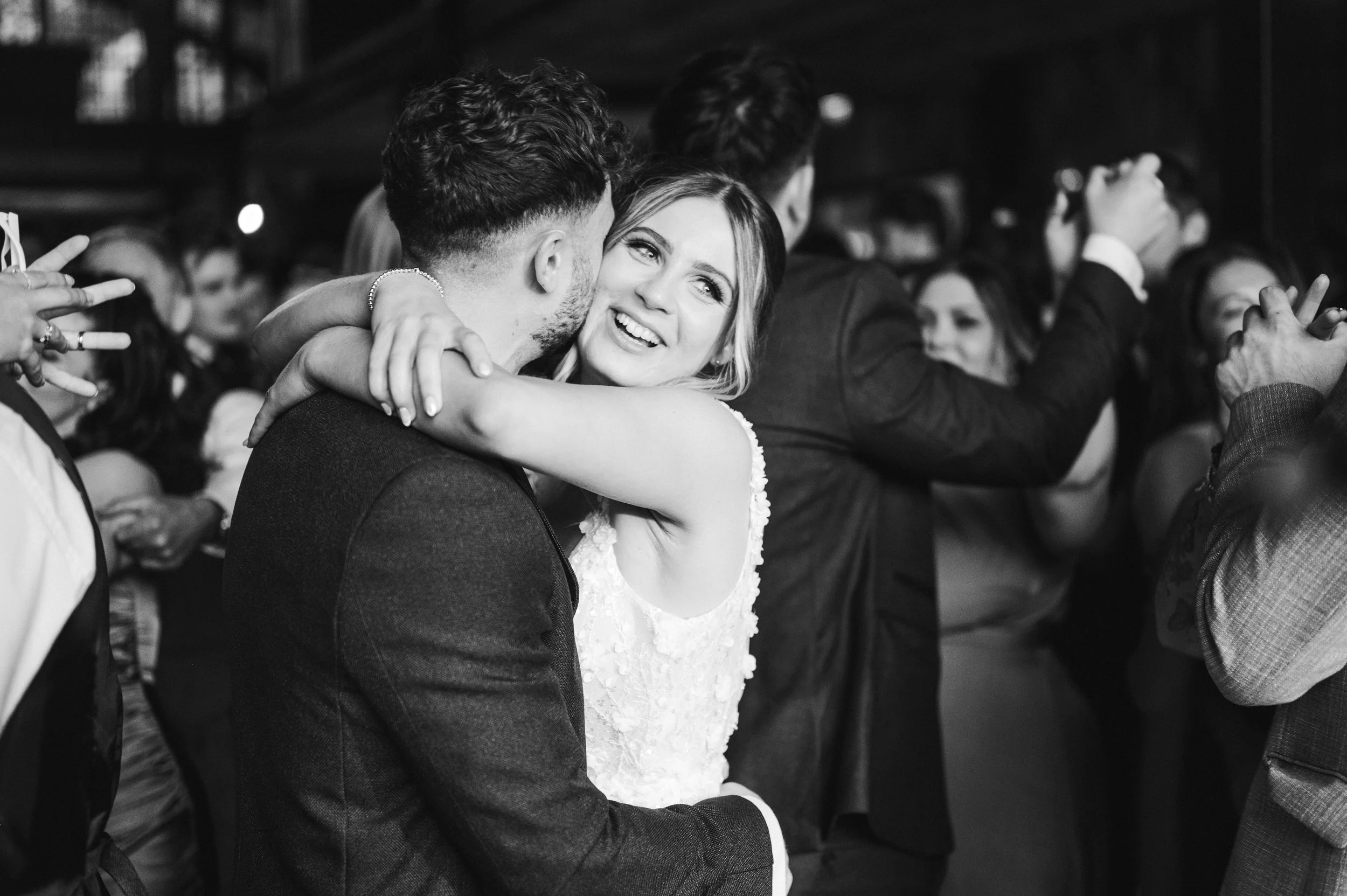 A black and white photo of a bride and groom dancing, with the bride holding the groom and smiling at him. The background shows other couples dancing at a wedding reception hidden river cabins, Gareth roy photography