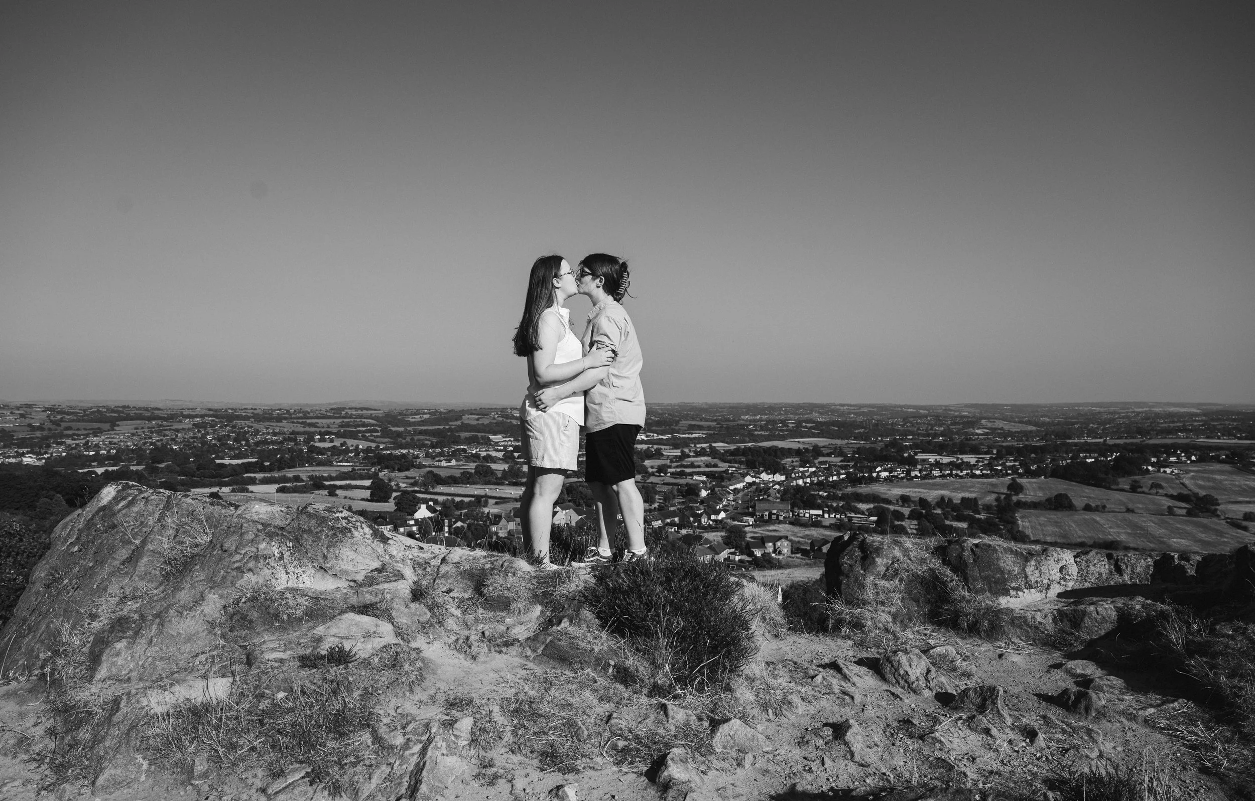 Mow Cop Engagement Shoot - Black and white image of two women stood in front of a landscape of Cheshire while embracing and kissing