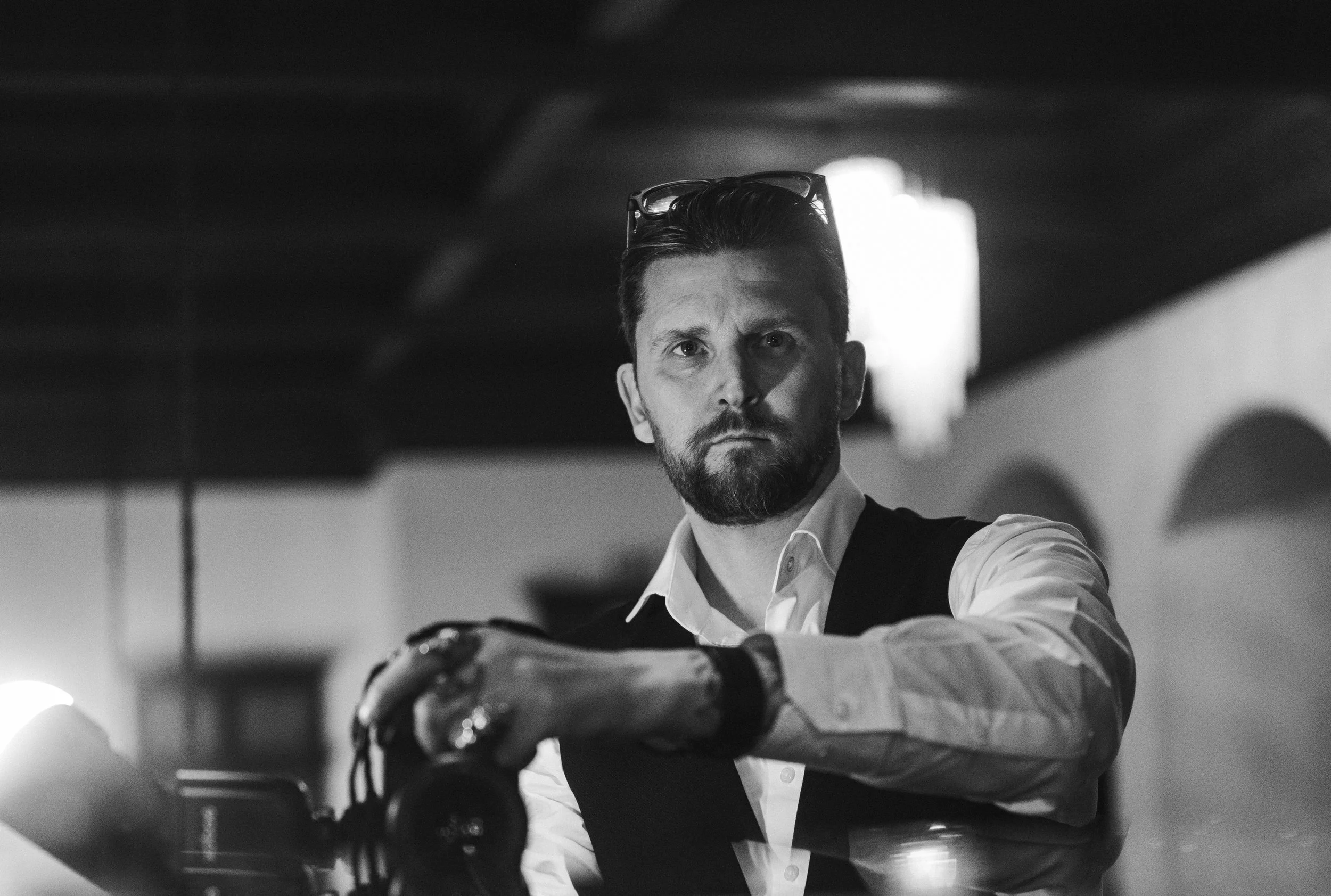 Black and white photo of a man with beard and short hair, wearing a white shirt with a vest, looking serious, with glasses resting on his head, sitting at a table with a camera in his hand