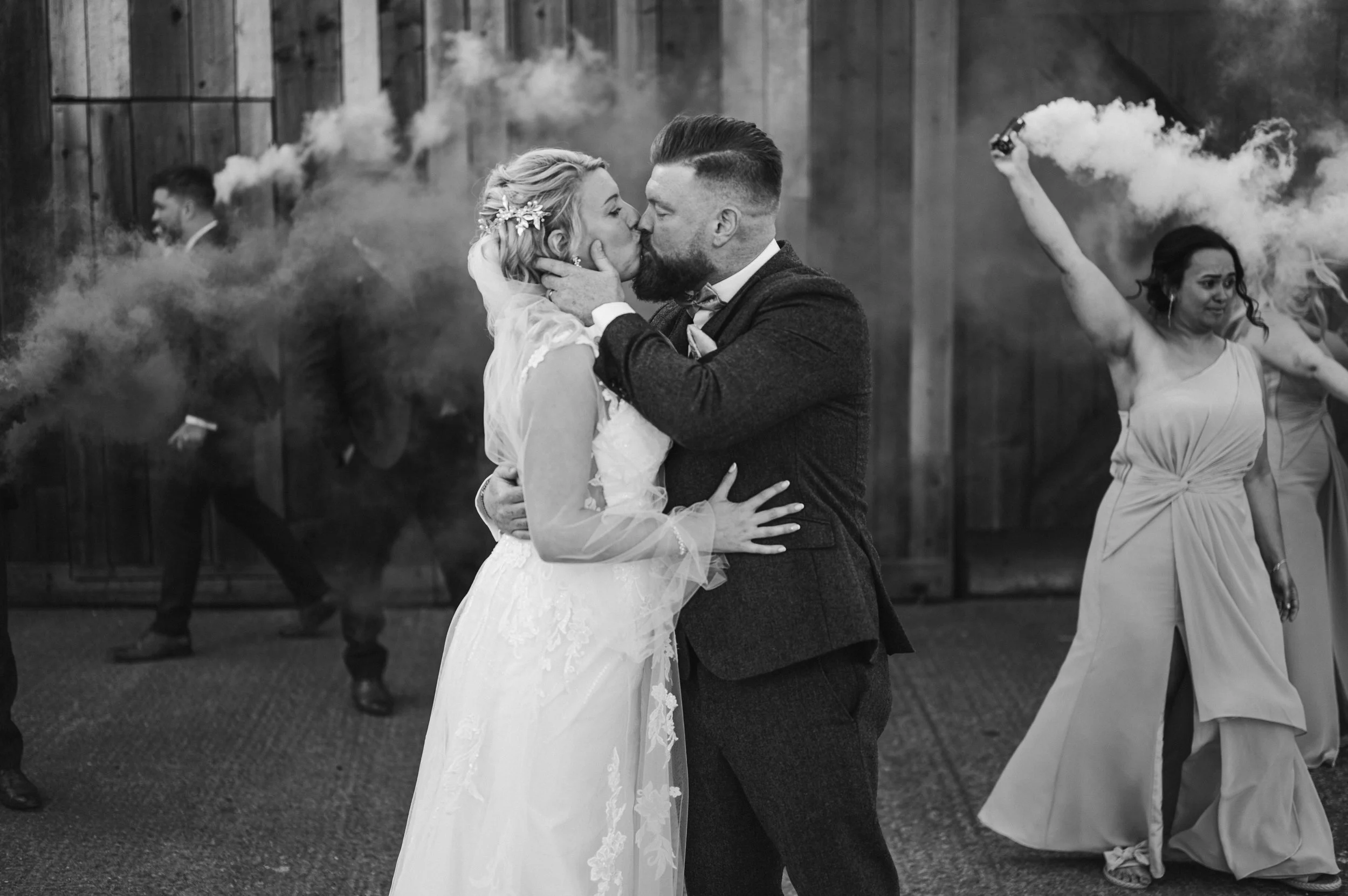 A black-and-white photo of a wedding, showing a couple kissing, with a smoky backdrop and bridesmaids dancing in the background grange barn Whitchurch, Gareth roy photography