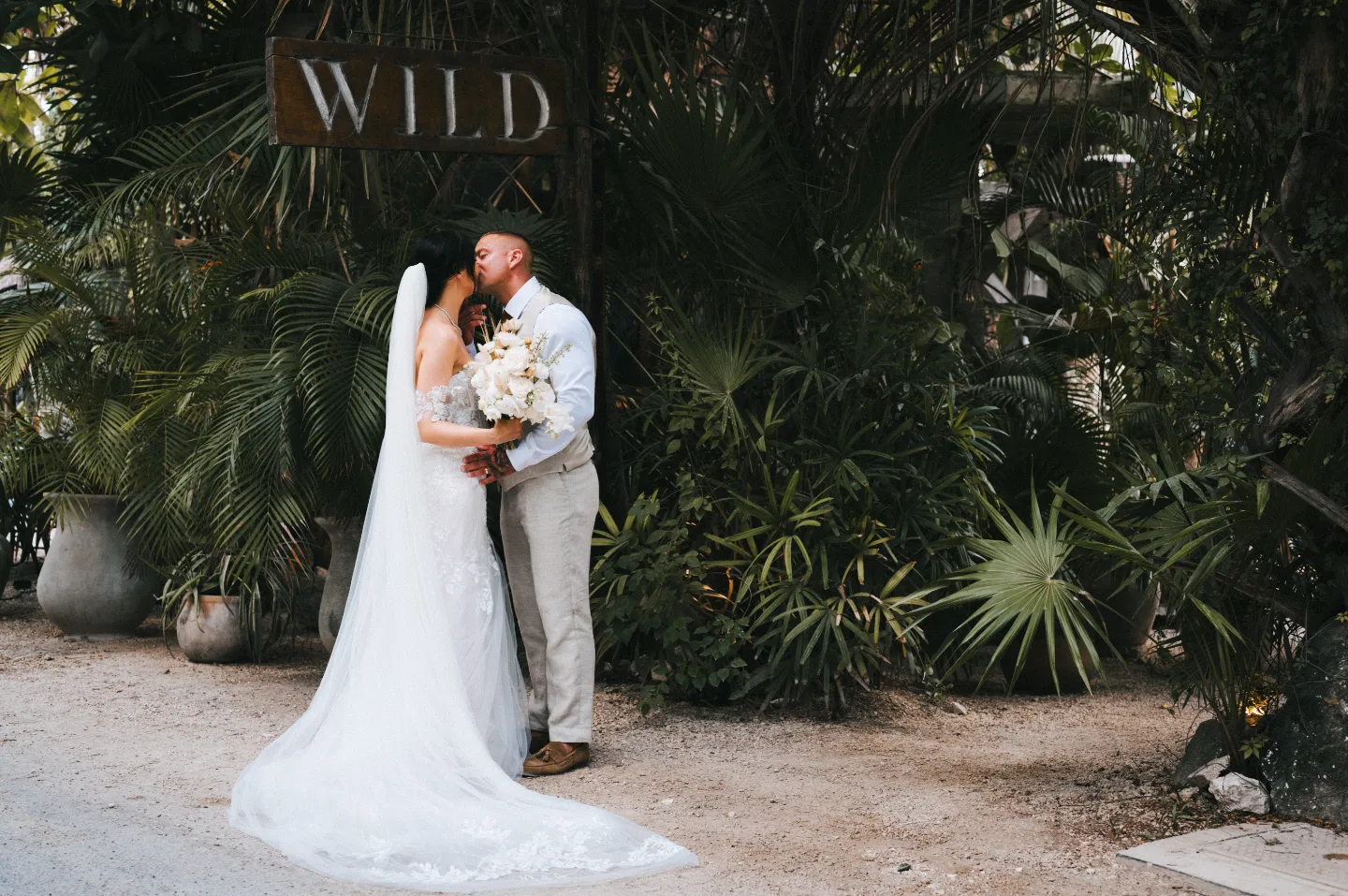 Bride and groom in wedding attire kissing in front of a background of greenery and a sign which says 'Wild'
