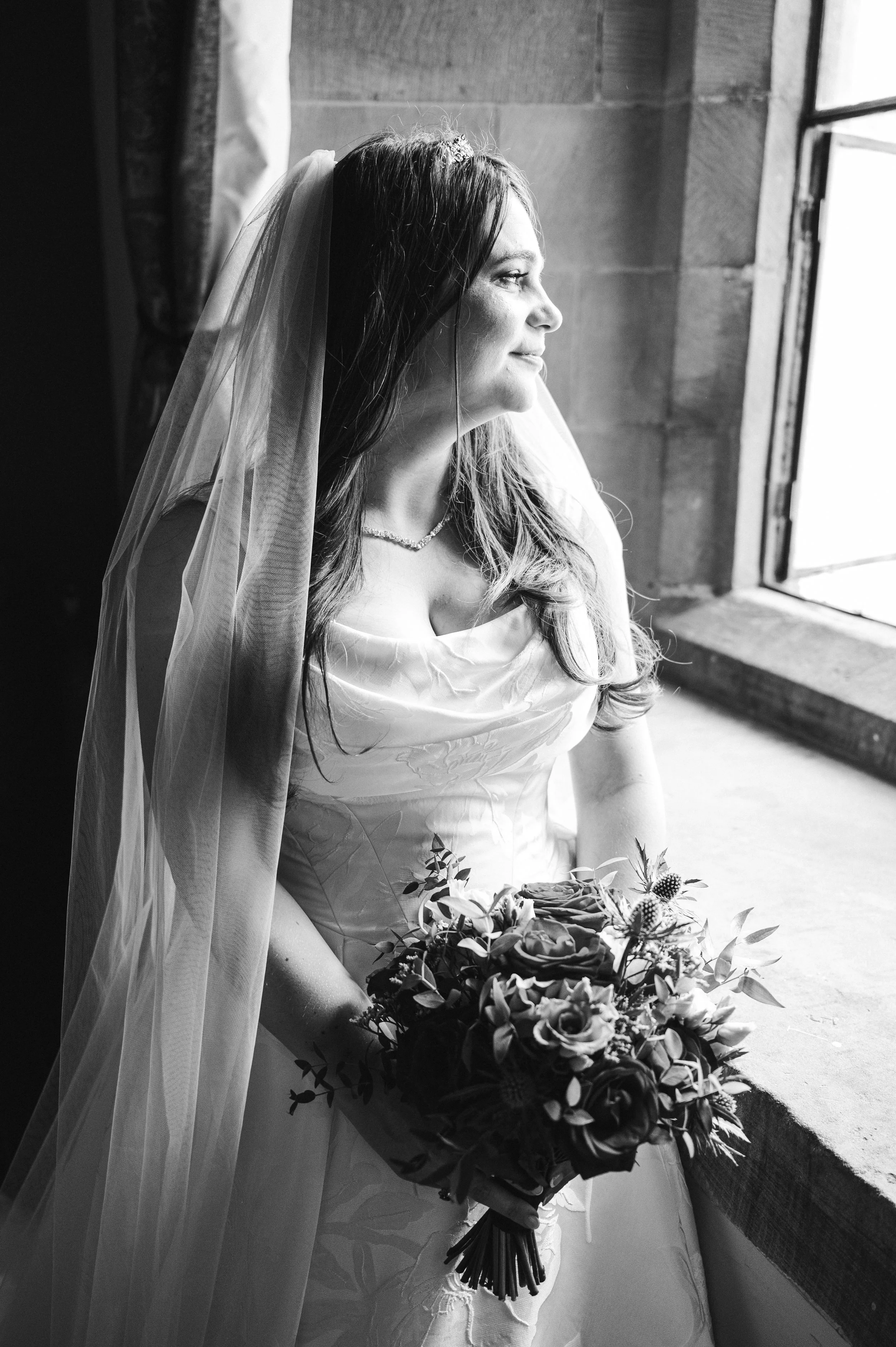 Peckforton Castle - black and white image of bride in wedding dress and holding bouquet while looking outside a bright window