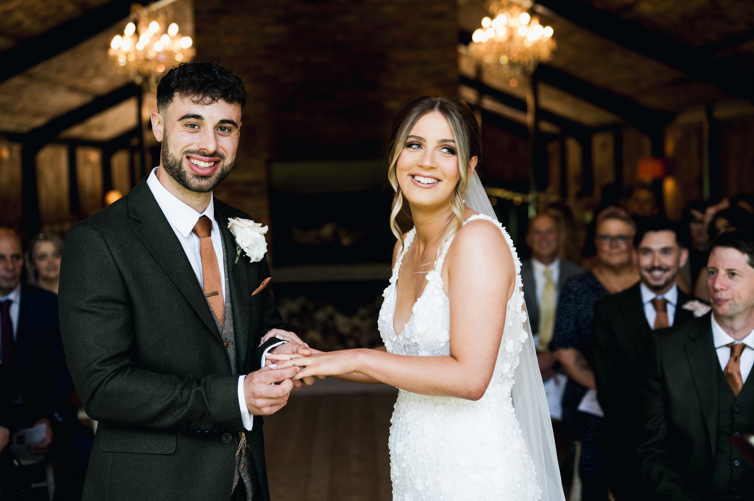 A bride and groom exchanging rings during their wedding ceremony in a warmly lit venue with chandeliers and friends in the background hidden river cabins, Gareth roy photography