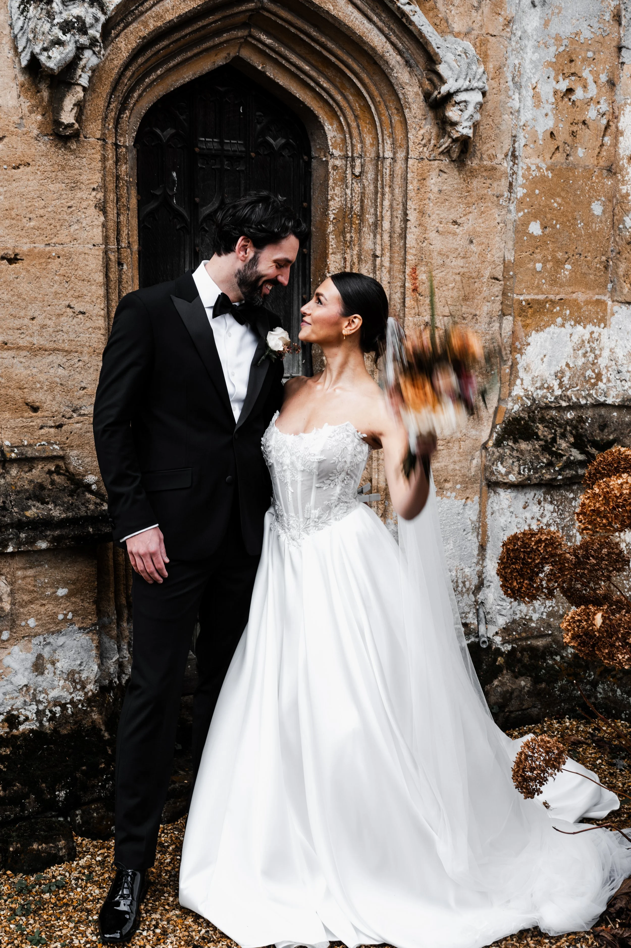 A bride and groom dressed in wedding attire stand close together outdoors near an old stone church, smiling and looking into each other's eyes; at sudeley castle, Gareth roy photography