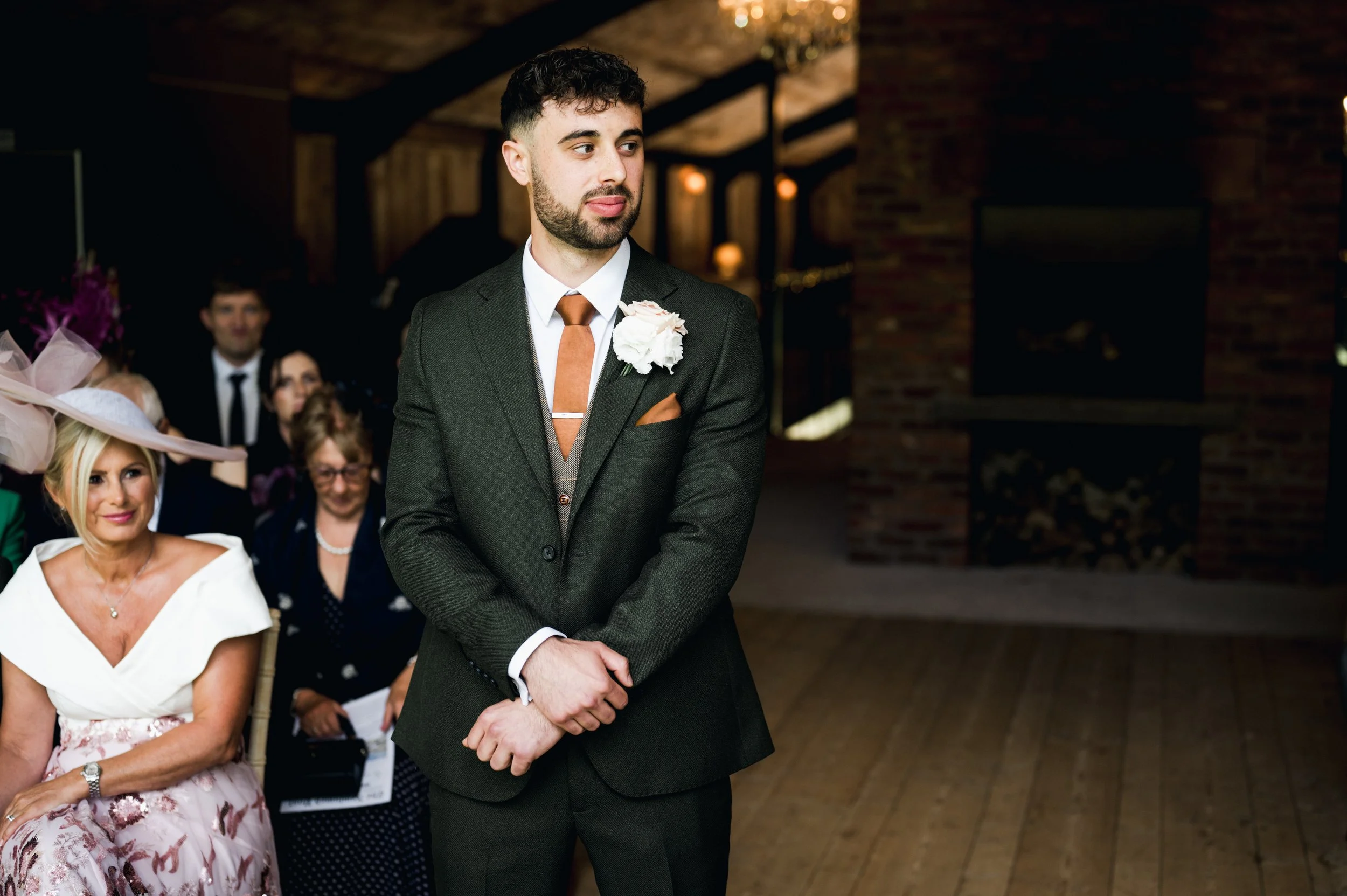 A groom in a dark suit with a white shirt and brown tie stands with his hands clasped, wearing a white boutonniere on his lapel hidden river cabins, Gareth roy photography