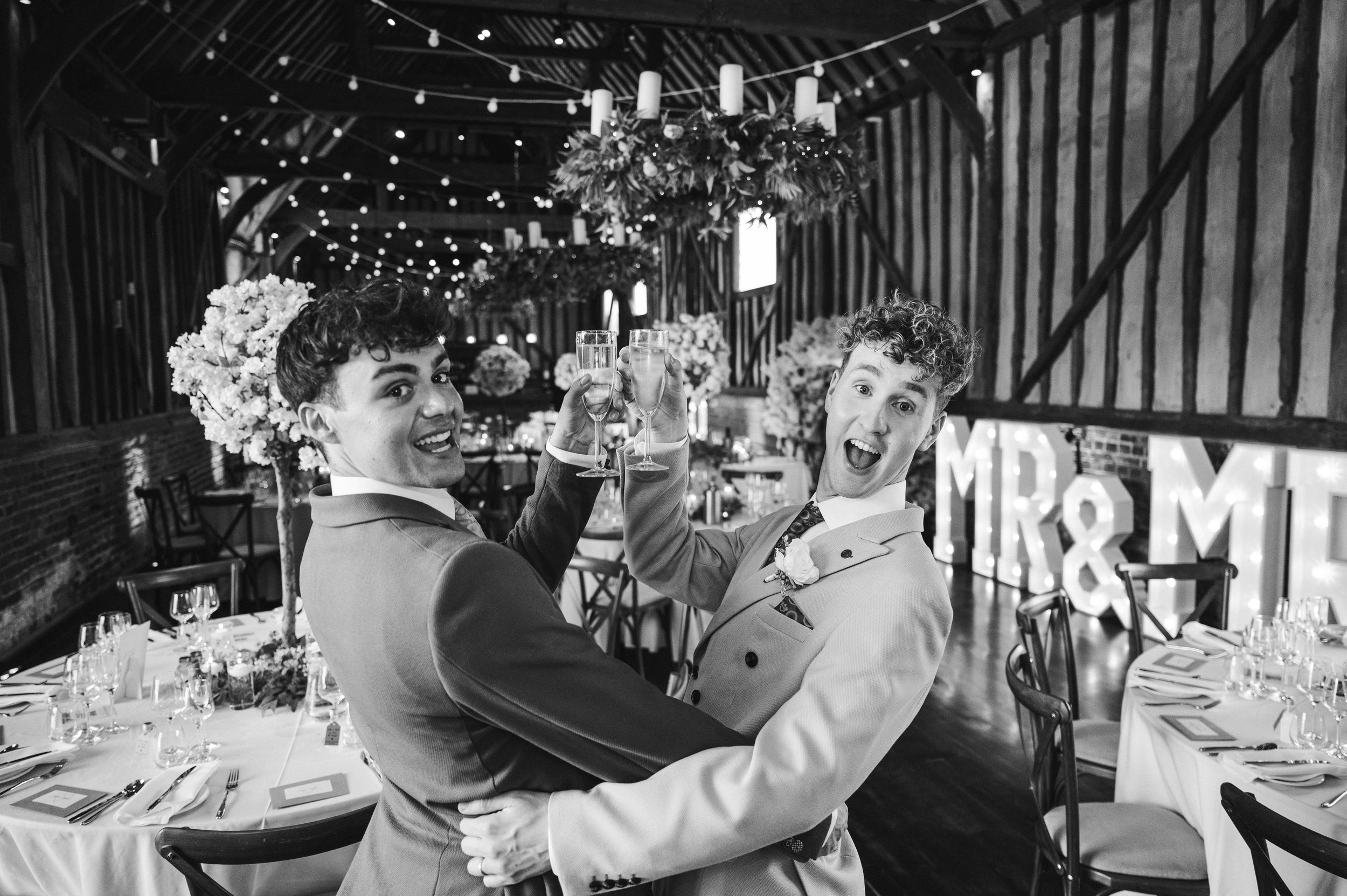 Black and white image of two grooms clinking glasses and looking back at the camera while the venue is behind them