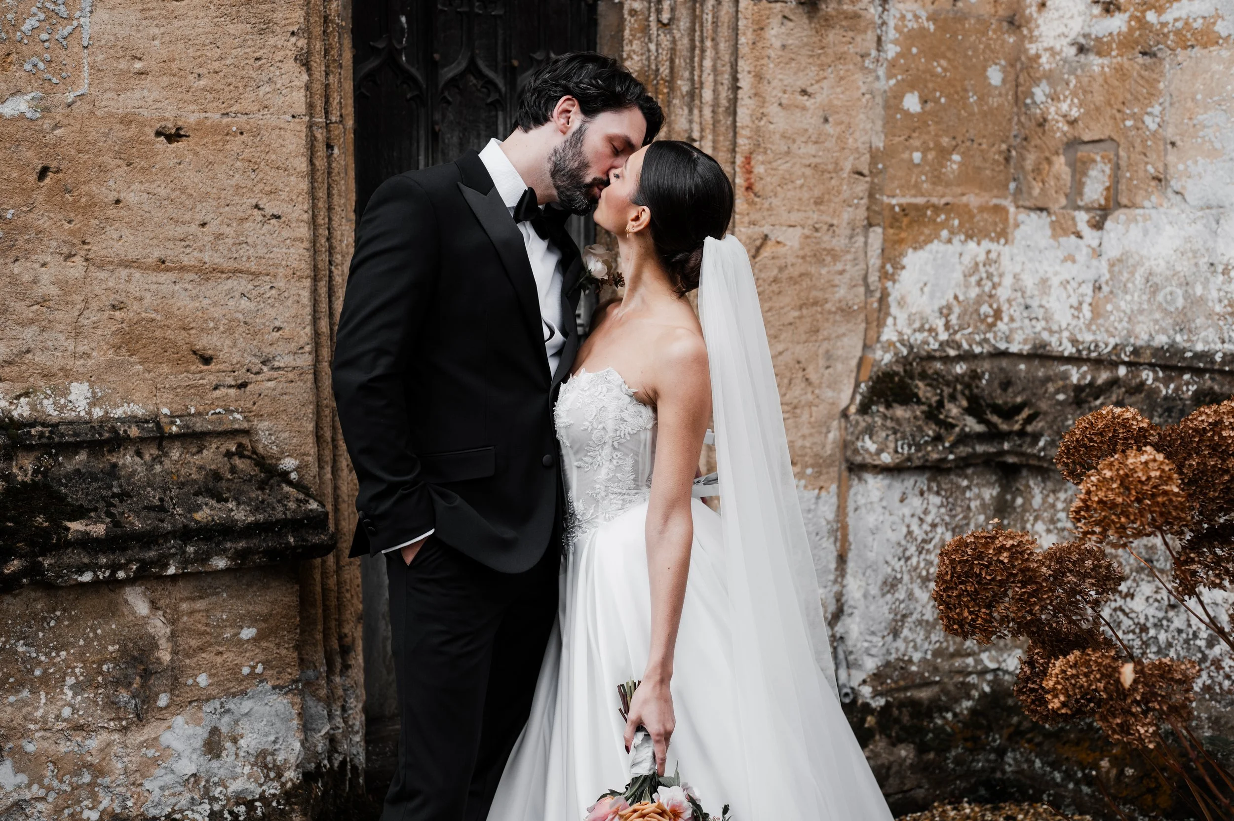 A bride and groom sharing a kiss outside, with vintage stone walls in the background. The bride is in a strapless white wedding dress and holding a bouquet, while the groom is in a black tuxedo at sudeley castle, Gareth roy photography