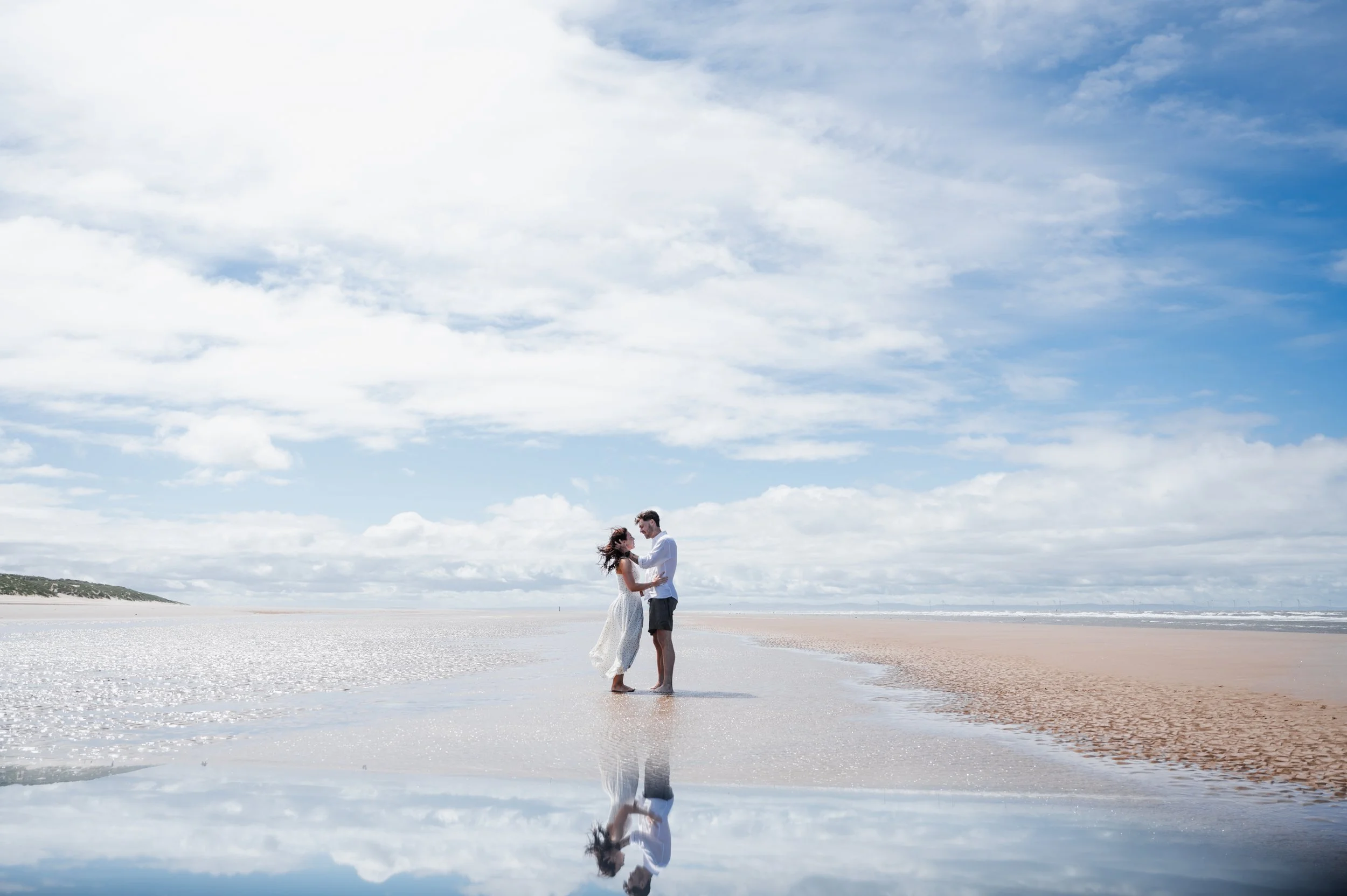 Formby Beach Engagement - Man and woman wearing white clothes stood in shallow water on a beach while embracing