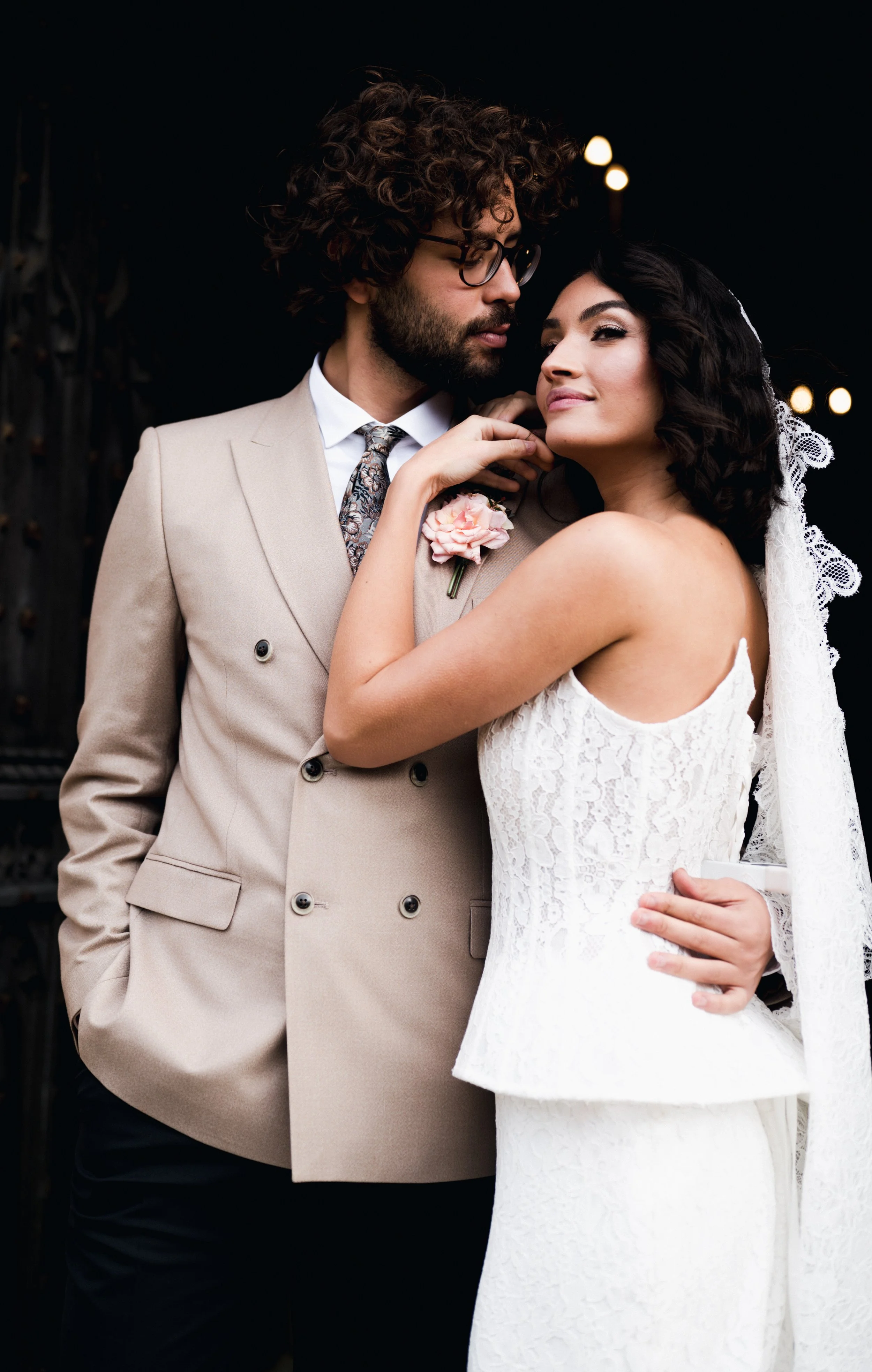A bride and groom embrace at their wedding, with the groom wearing a beige suit and floral tie, and the bride in a white lace wedding dress with a veil at sudeley castle