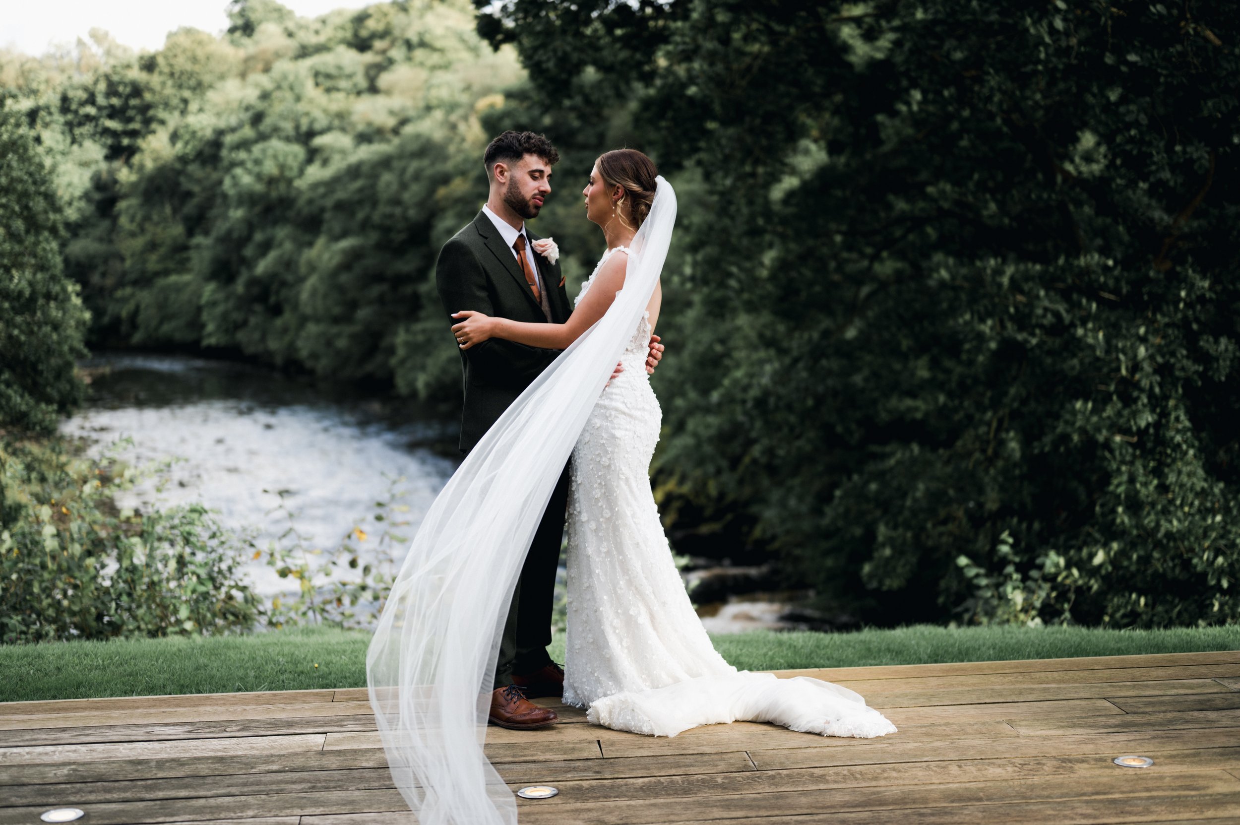 A bride and groom standing on a wooden platform outdoors, embracing each other, with trees and a river in the background hidden river cabins, Gareth roy photography