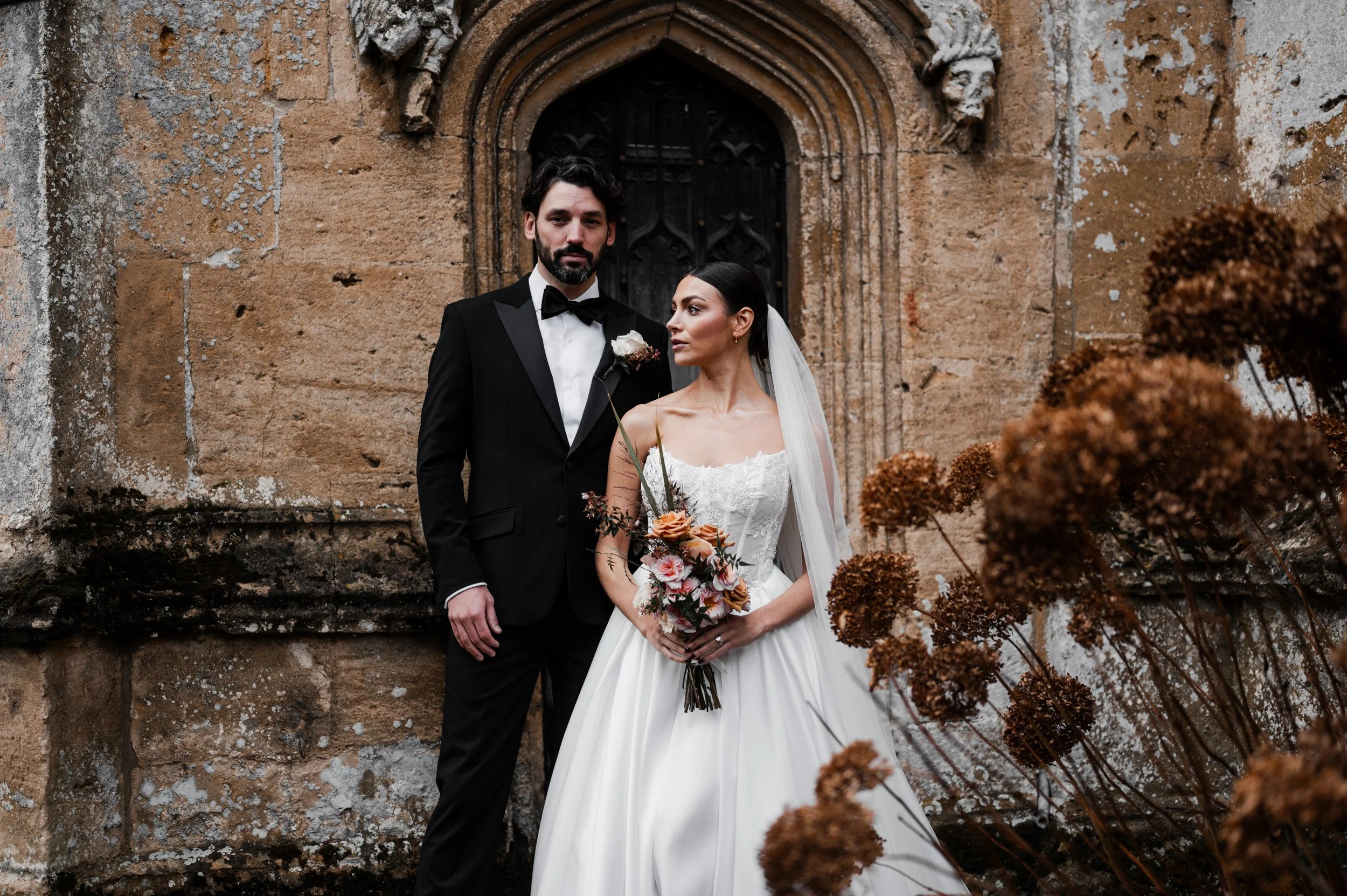 A bride and groom standing outside an old stone building, with the bride holding a bouquet of flowers and the groom dressed in a black tuxedo at sudeley castle, Gareth roy photography