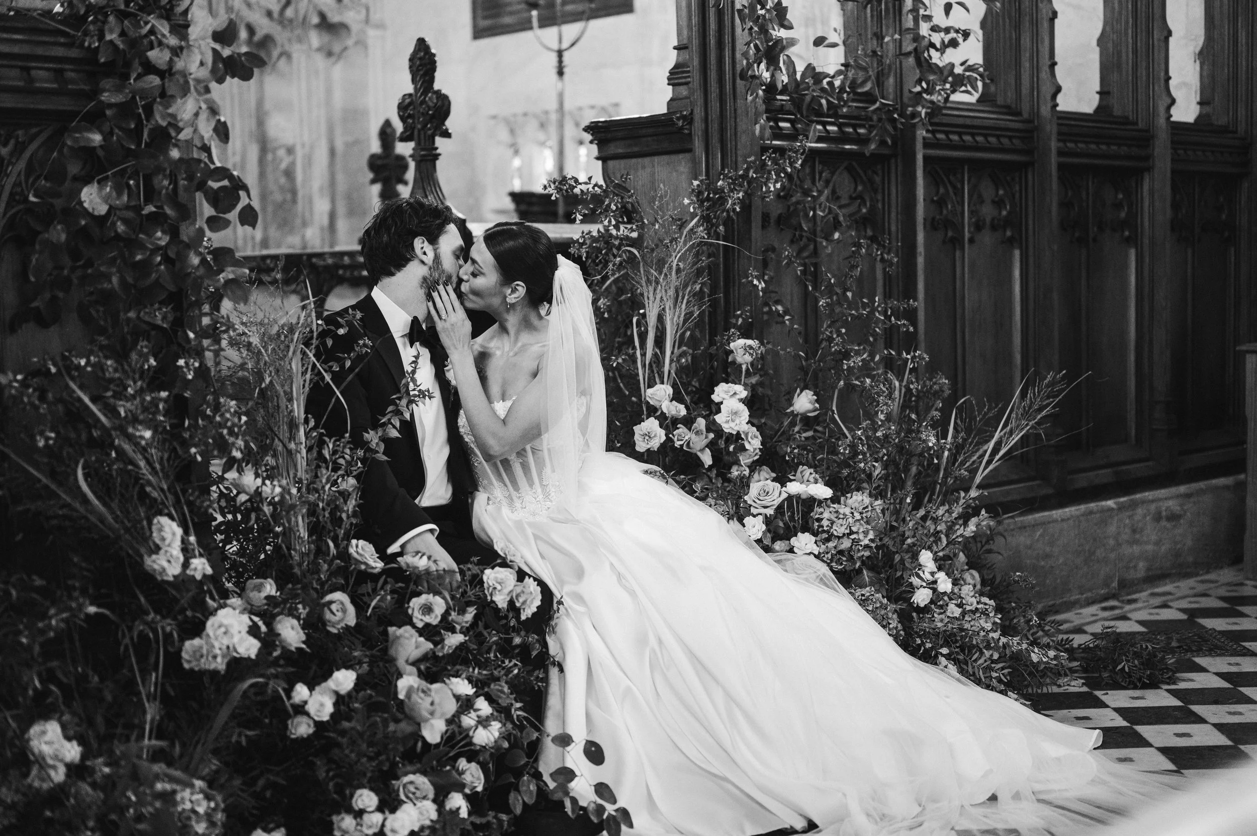 Black and white photo of a bride and groom sitting close together, surrounded by flowers and plants, in a church setting at sudeley castle, Gareth roy photography