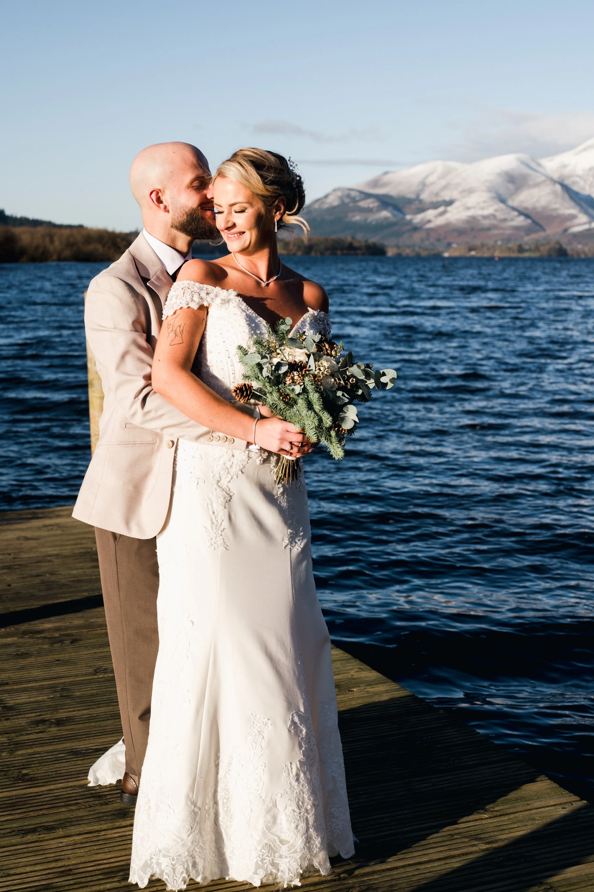 Bride and groom stood on a pier over a lake with snowy mountains behind. 
