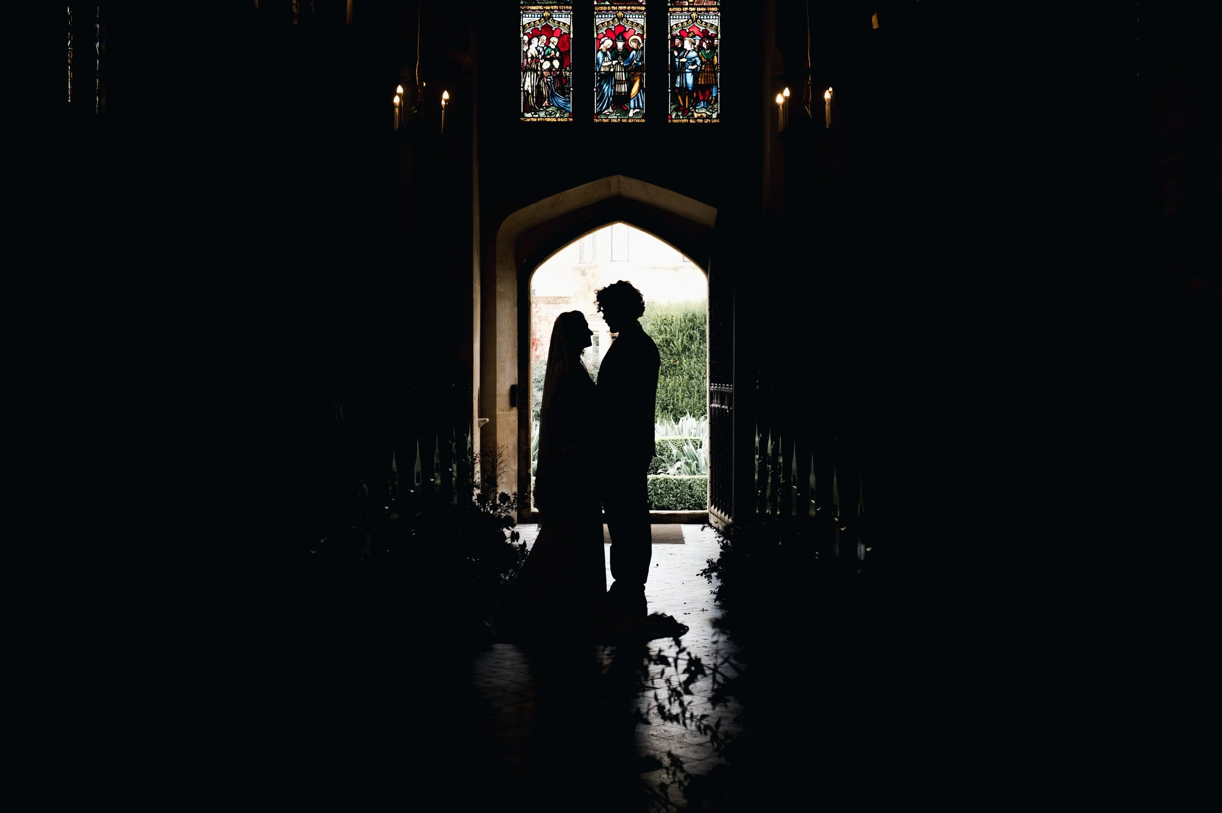 Wedding Photography Cheshire - Silhouettes of bride and groom stood in the entrance of a church doorway with stained glass windows above. 