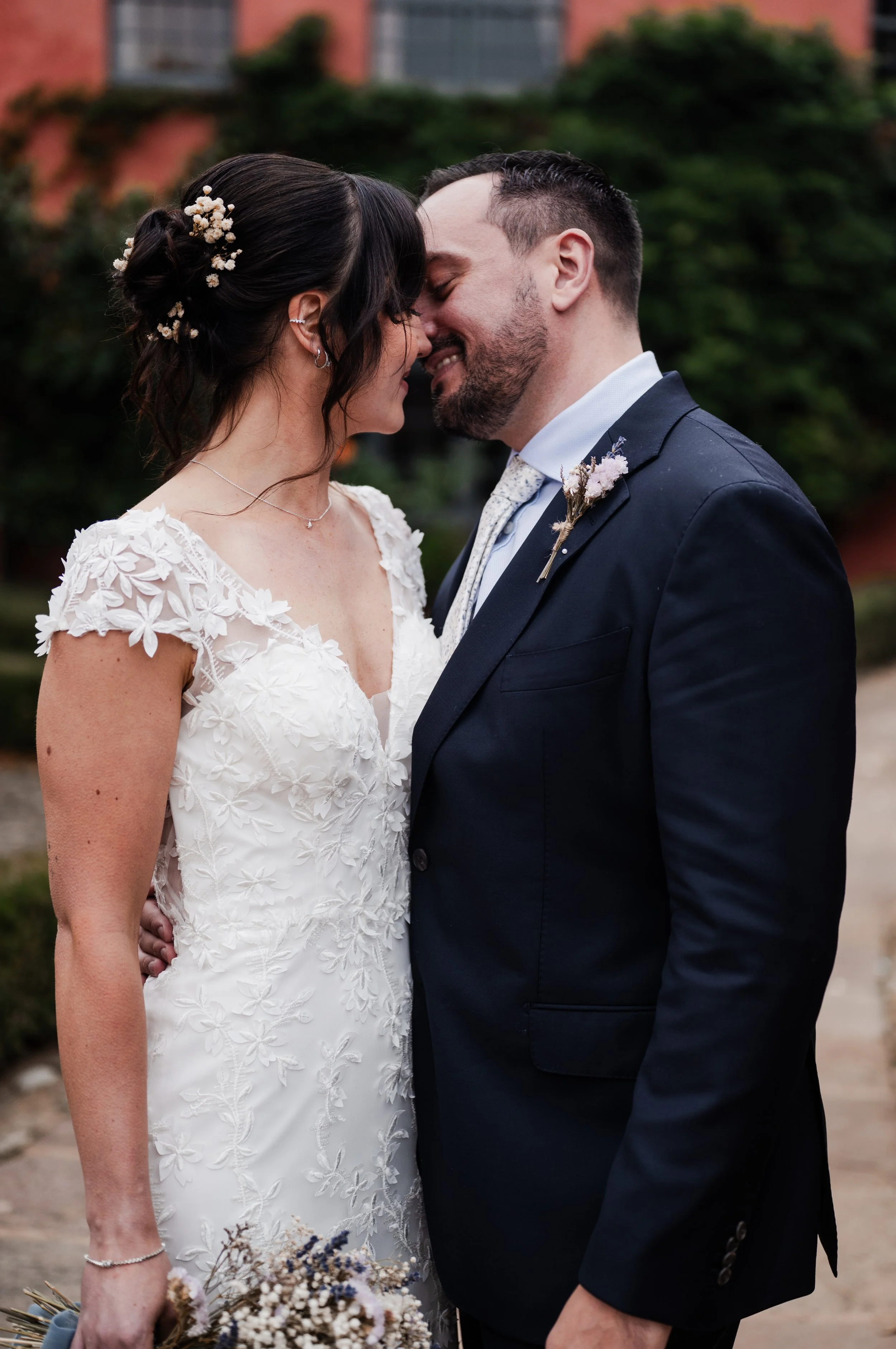 Man and a woman in wedding attire, woman is in a white lace dress and man is in a dark suit and they are standing close together.