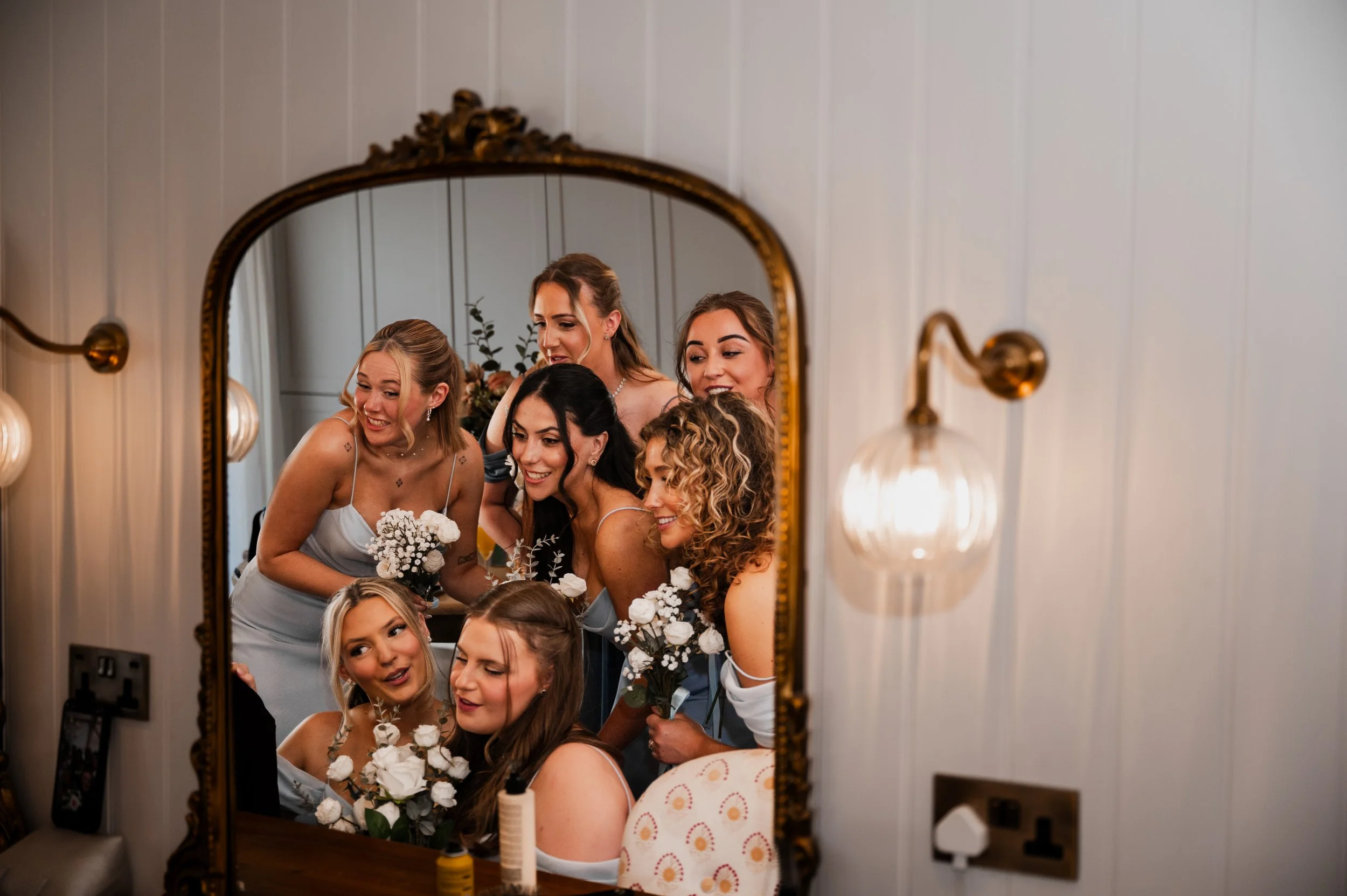 A group of seven bridesmaids sit in front of a mirror dressed in light blue dresses and holding small white bouquets
