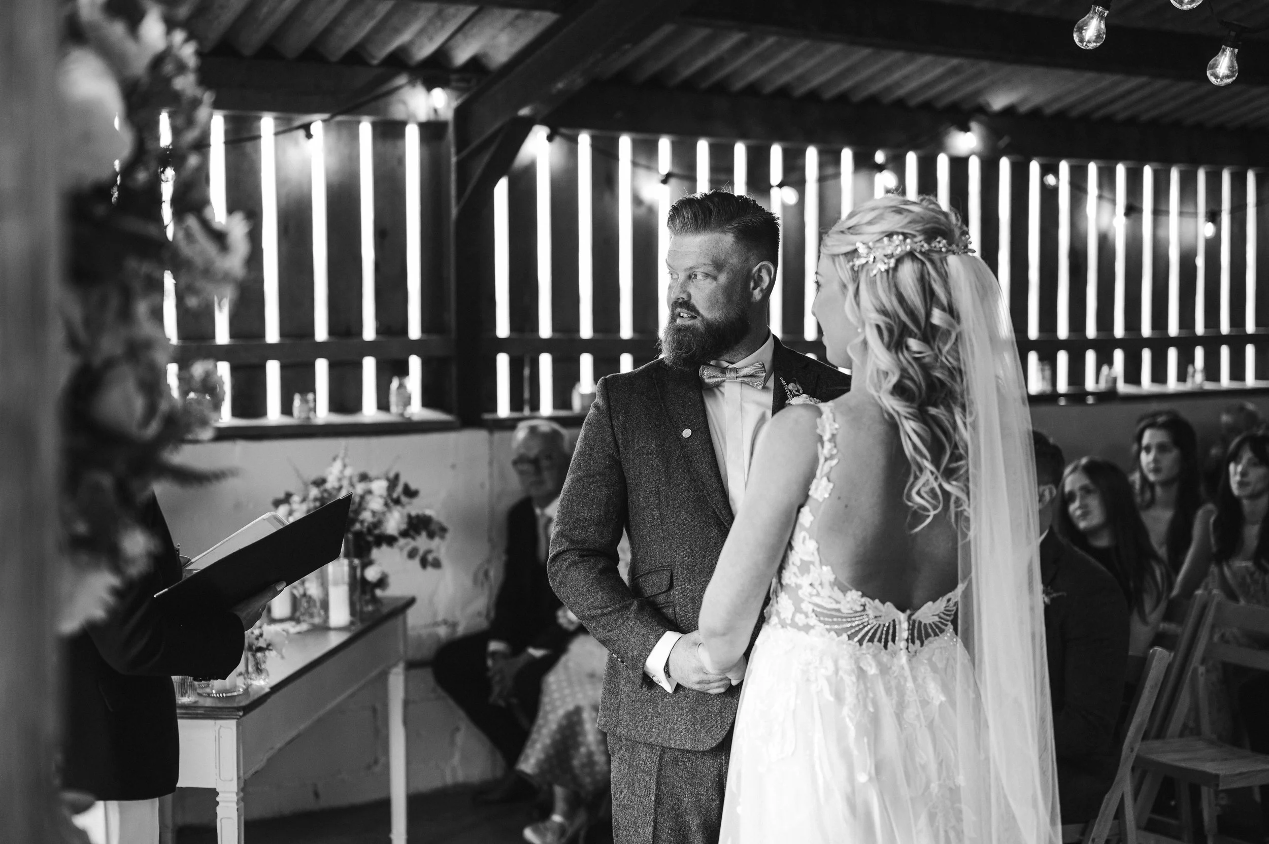 A black and white photo of a wedding ceremony with a bride and groom standing together. The bride has blonde hair with a floral headpiece, wearing a lace wedding gown grange barn Whitchurch, Gareth roy photography
