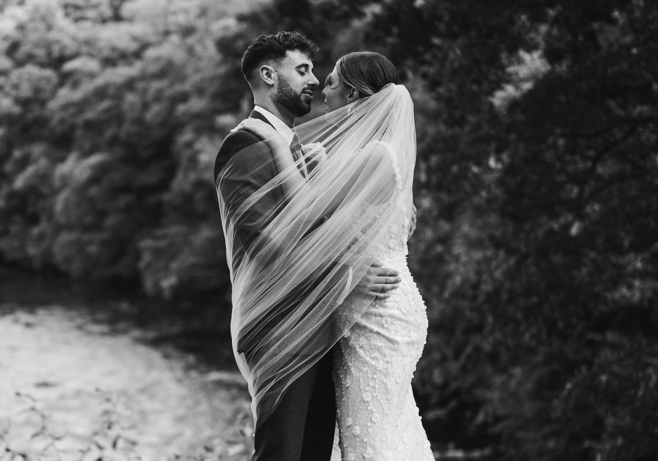 Black and white photo of a newlywed couple; the groom is kissing the bride on the forehead, and they are smiling with their eyes closed, standing against a brick wall. The bride is holding a floral bouquet.