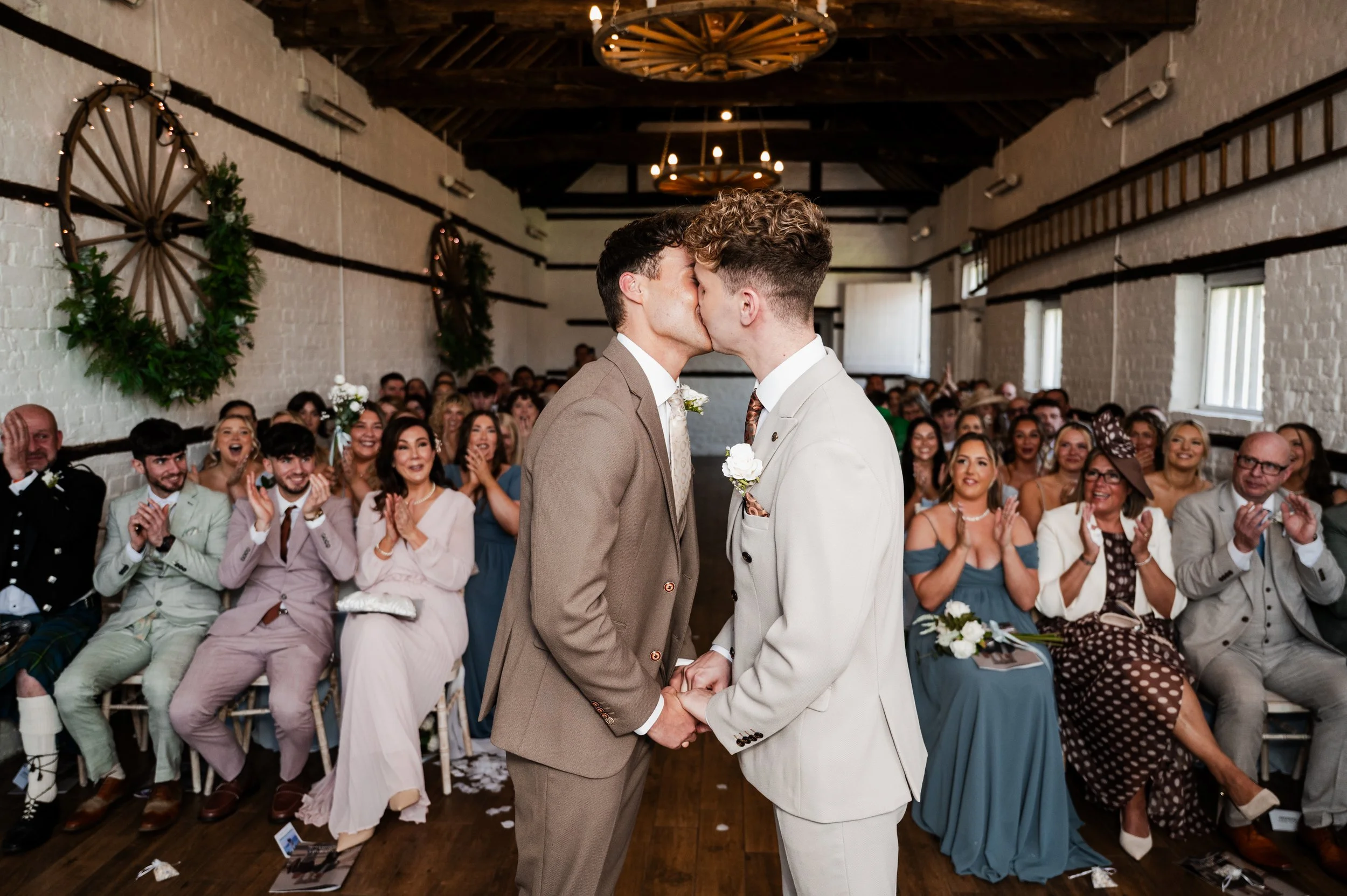 Two grooms kiss at the end of a wedding ceremony with all their friends and family clapping and smiling behind them
