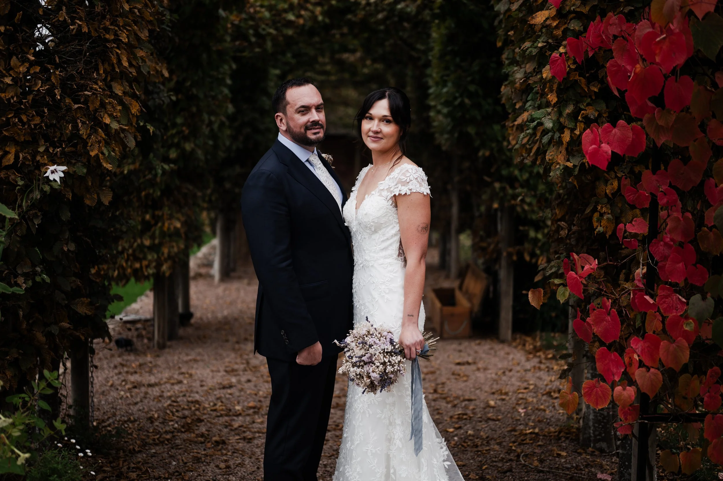 A bride and groom standing outdoors in a garden archway, dressed in wedding attire, looking at the camera pauntley court, Gareth roy photography