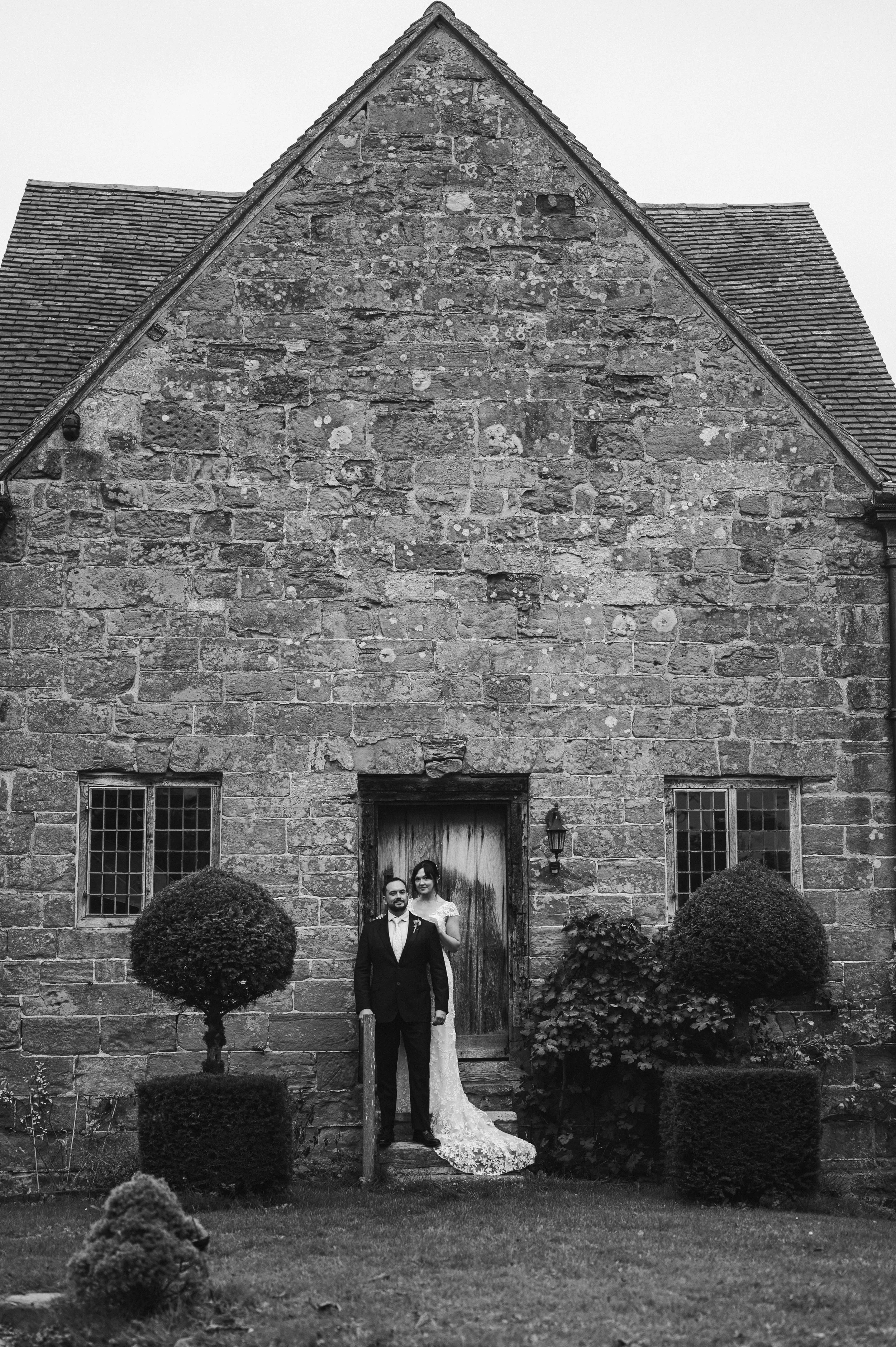 A black and white photo of a bride and groom standing in front of an old stone building with a wooden door at sudeley castle, Gareth roy photography