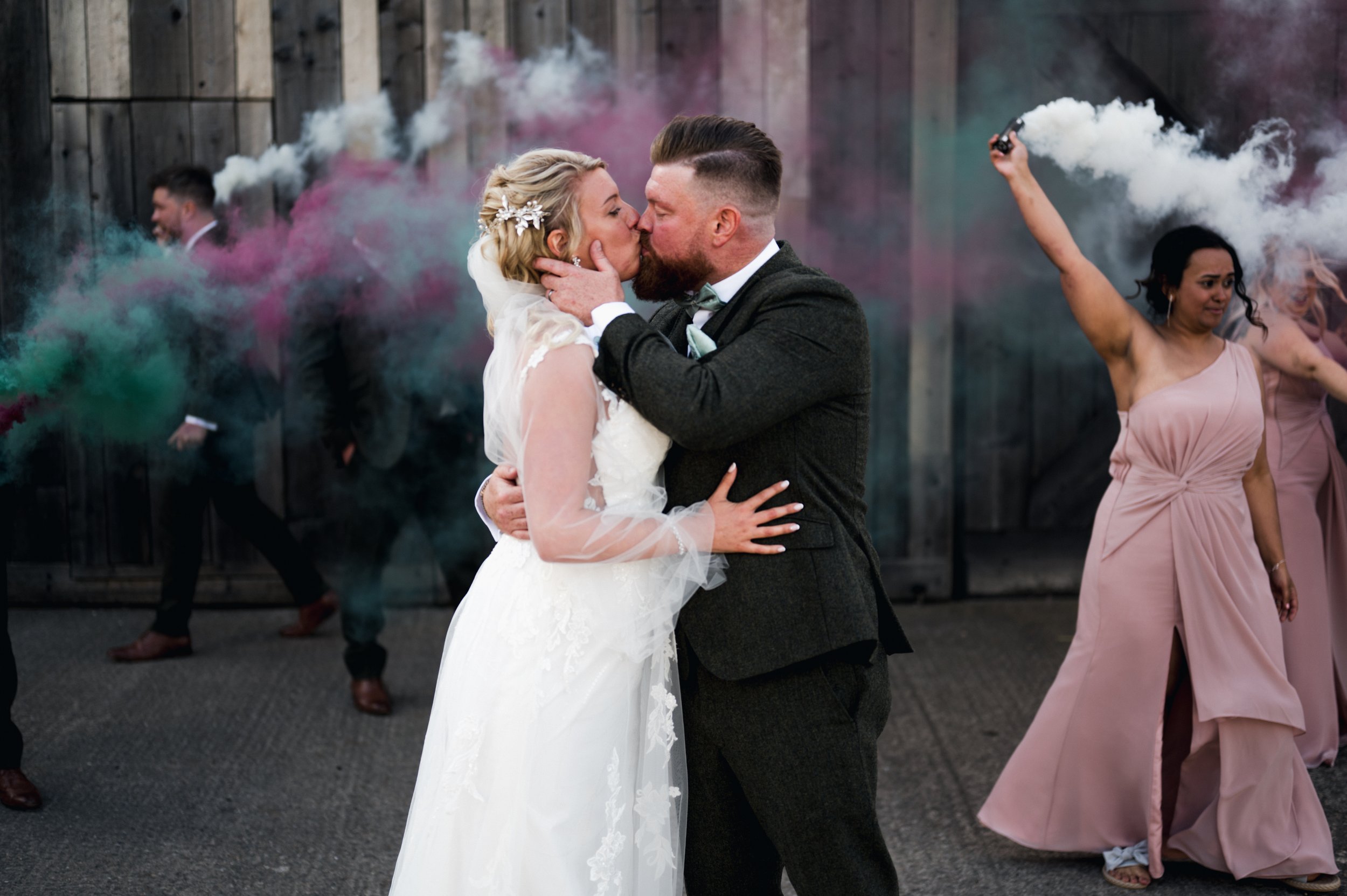 Bride and groom kissing at their wedding, with women in pink dresses and smoke bombs releasing colorful smoke in the background grange barn Whitchurch, Gareth roy photography