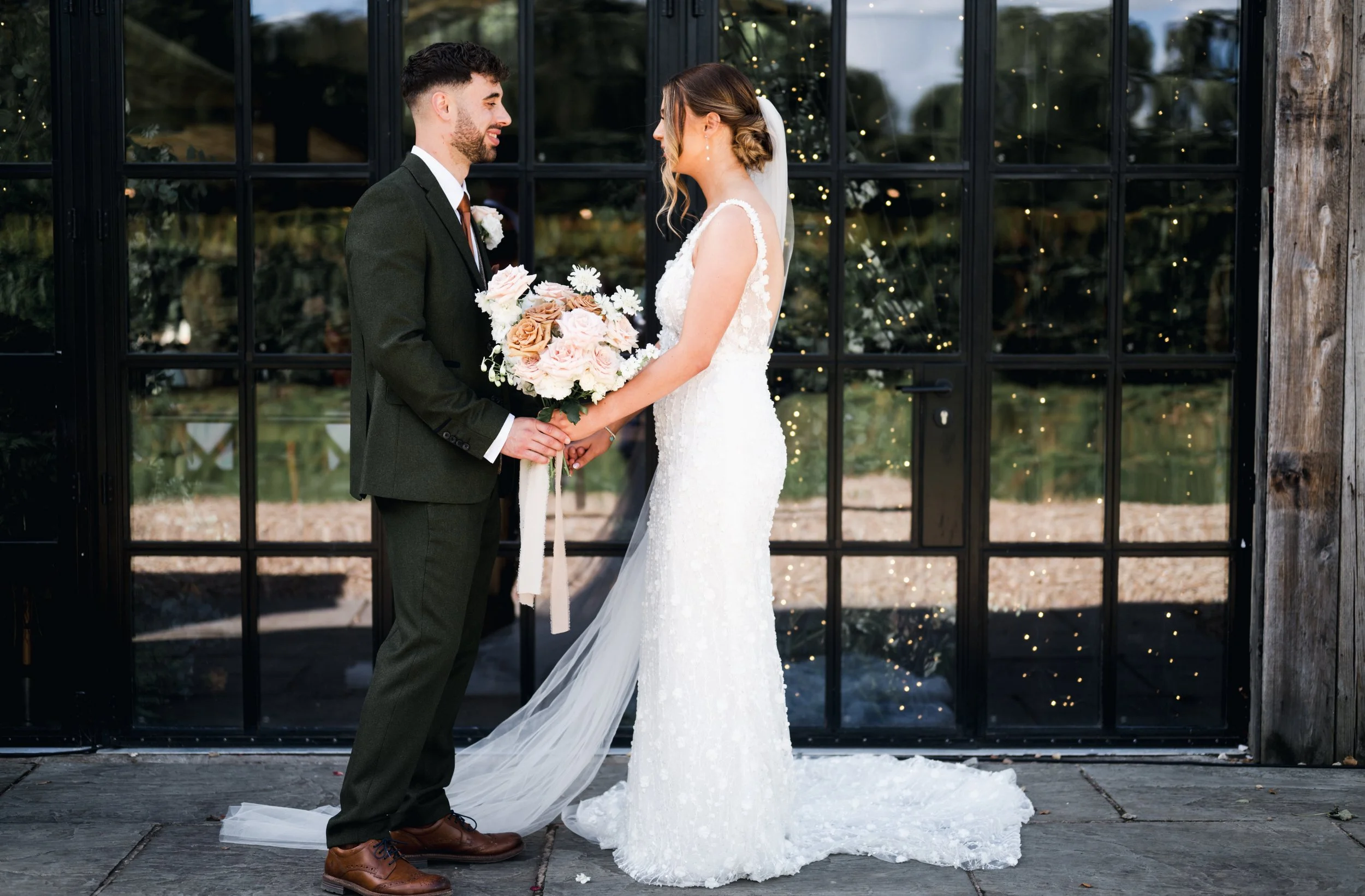 A bride and groom holding hands and facing each other during their wedding ceremony, standing outdoors in front of a black-framed glass door hidden river cabins, Gareth roy photography