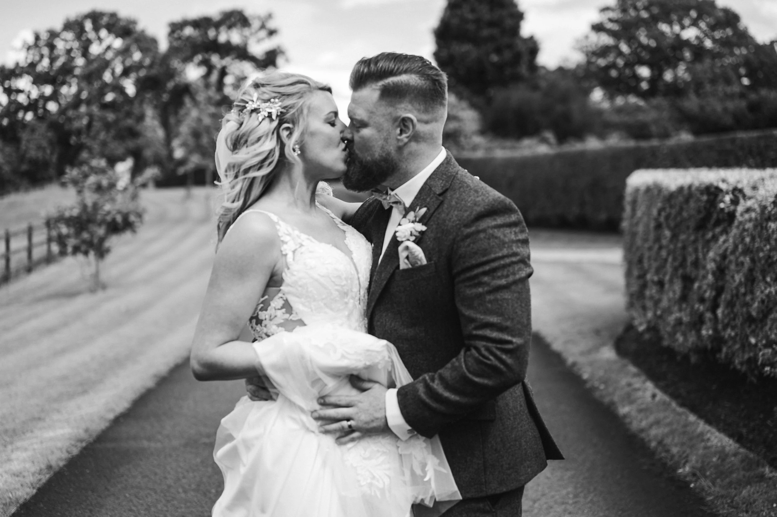 Black-and-white photo of a bride in an wedding dress holding a bouquet of flowers and a groom in a tuxedo with a bow tie, standing outside a stone building with an arched door.