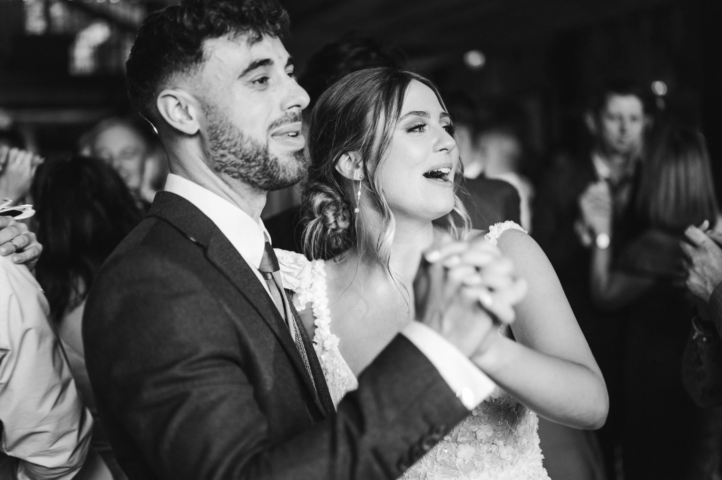 A black and white photo of a man and woman at a wedding, with the woman in a lace wedding dress and the man in a suit, both singing or reciting during the ceremony, surrounded by other guests hidden river cabins, Gareth roy photography