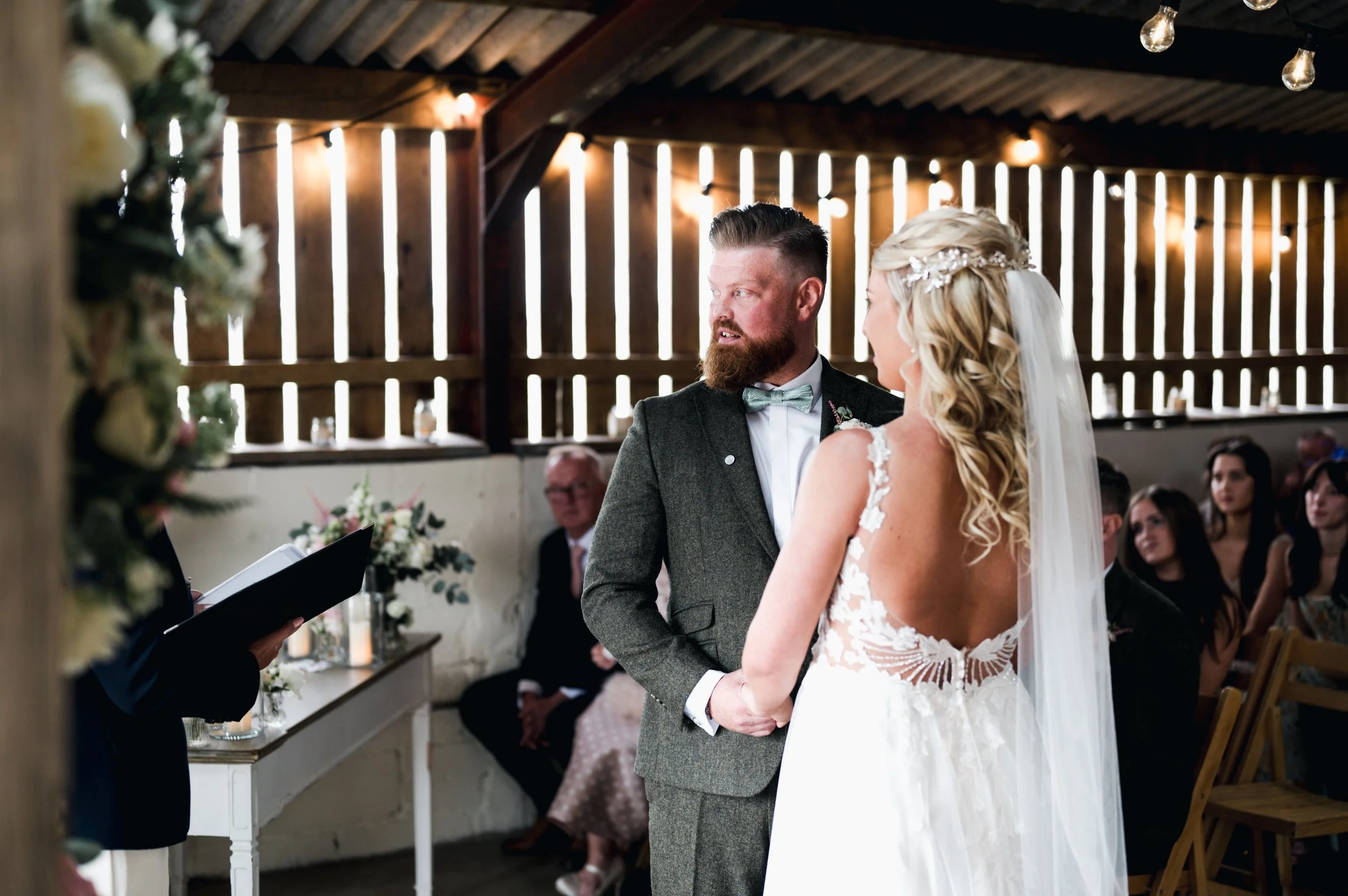 Bride and groom exchanging vows at an indoor wedding ceremony, with guests watching in the background grange barn Whitchurch, Gareth roy photography