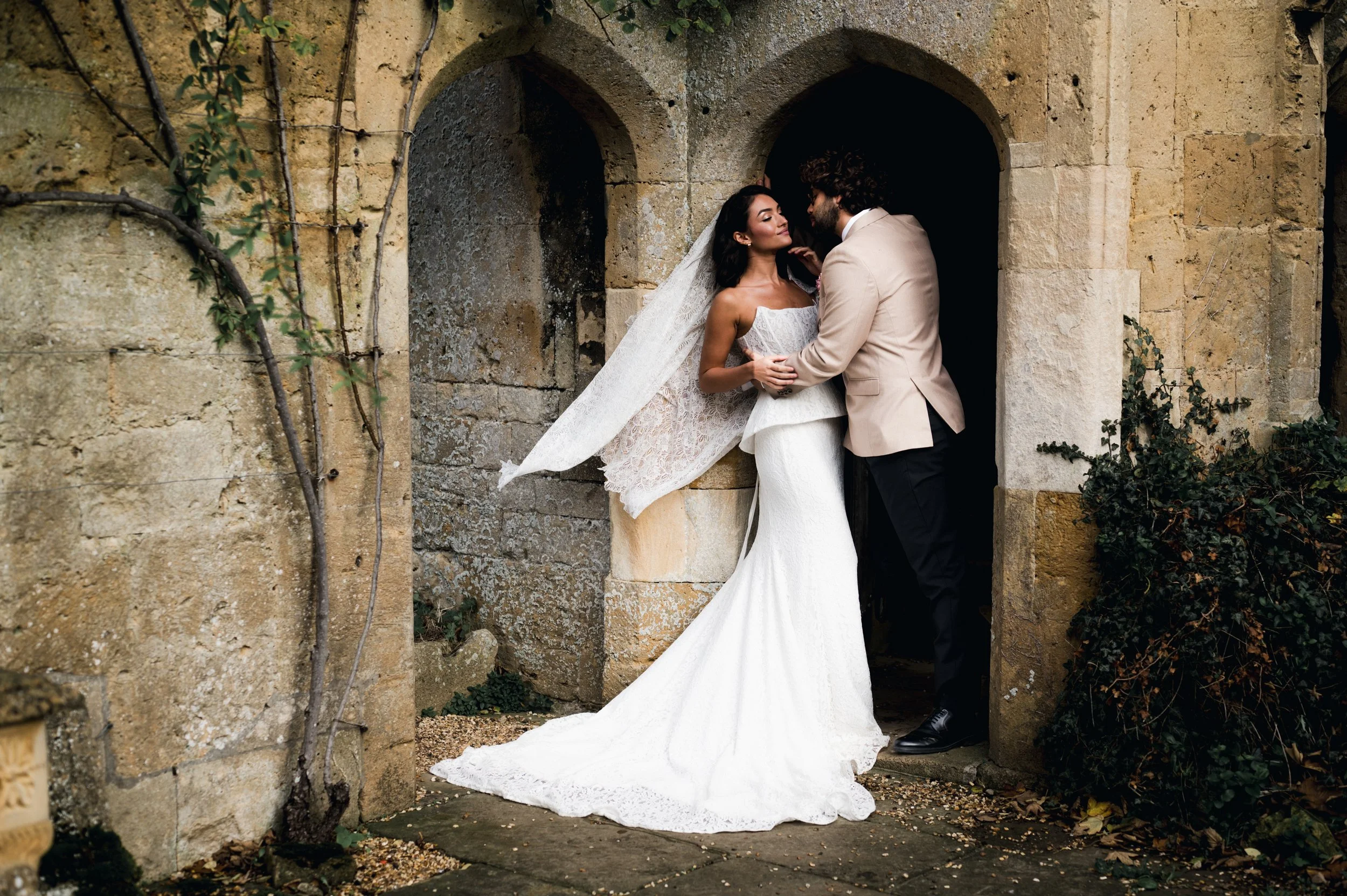Bride and groom embracing in a stone archway, the bride in a white lace wedding gown with a long train and veil, the groom in a beige suit jacket and dark pants, both gazing at each other at sudeley castle, Gareth roy photography