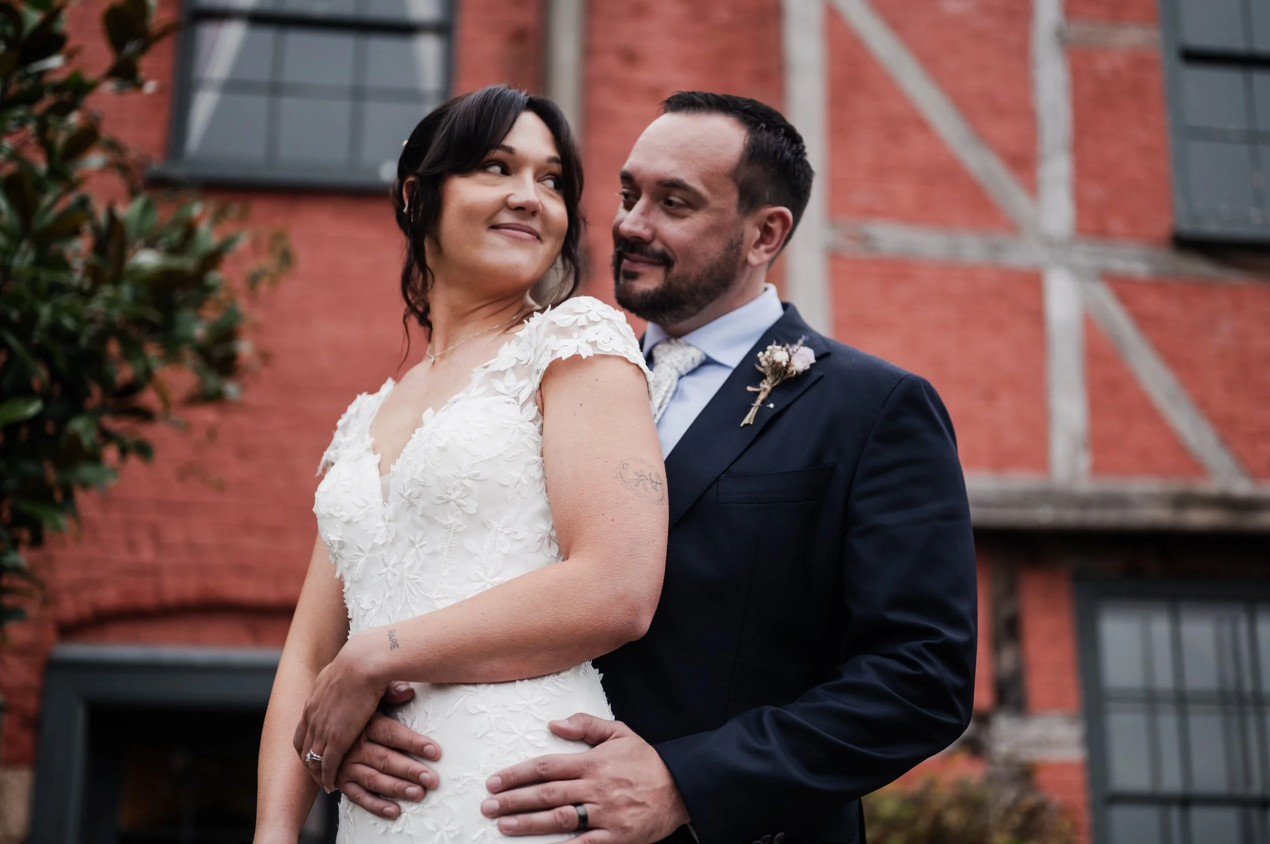 A bride and groom stand close together outdoors in front of a brick building, smiling as they look at each other pauntley court, Gareth roy photography