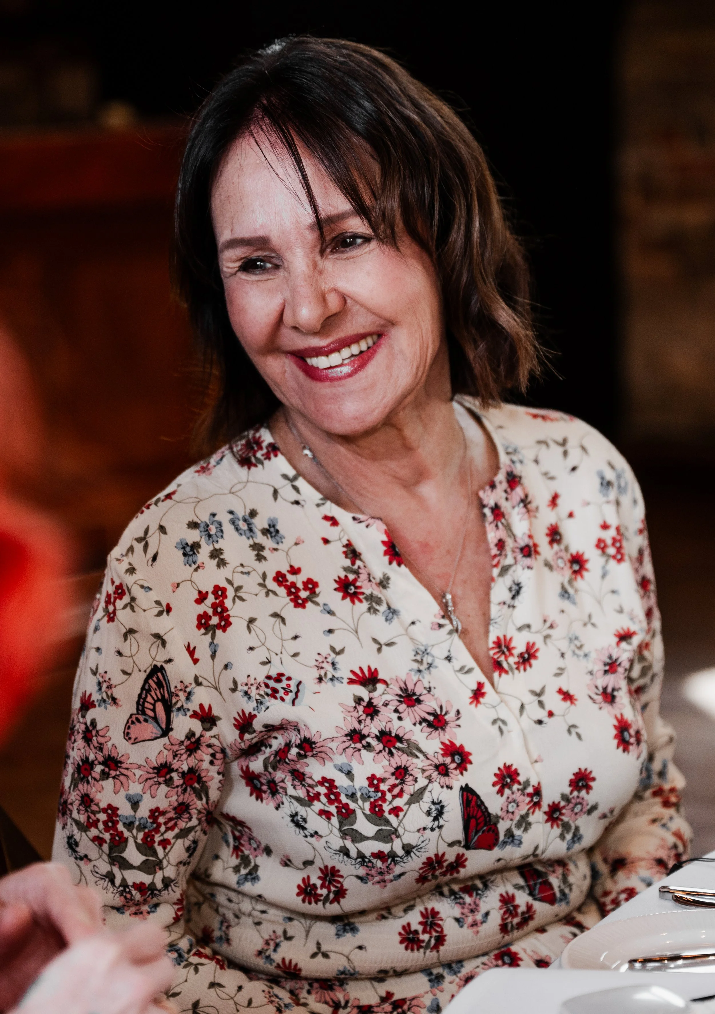Woman sitting at a table and smiling wearing a floral dress with butterflies on