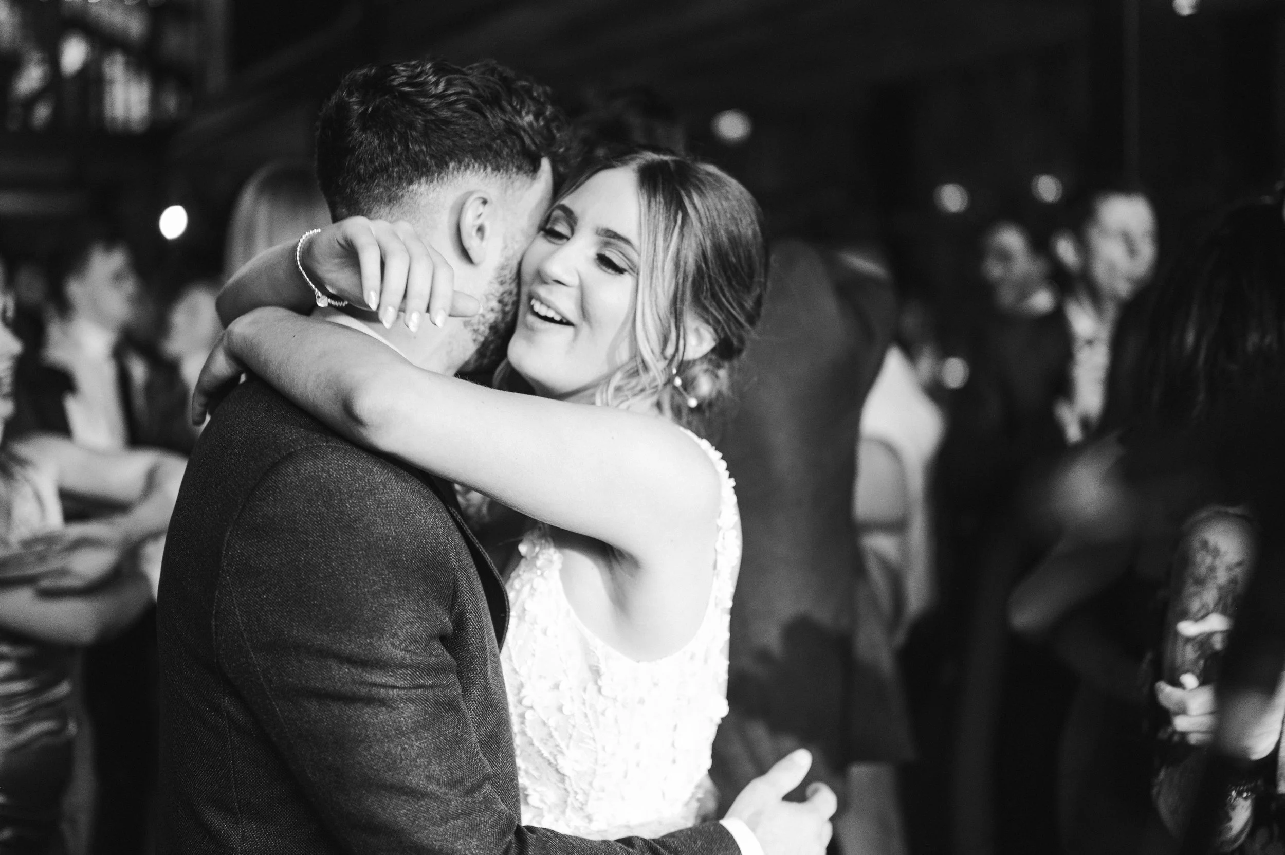 A woman in a wedding dress embraces a man in a suit, smiling happily, at a wedding reception with guests in the background hidden river cabins, Gareth roy photography