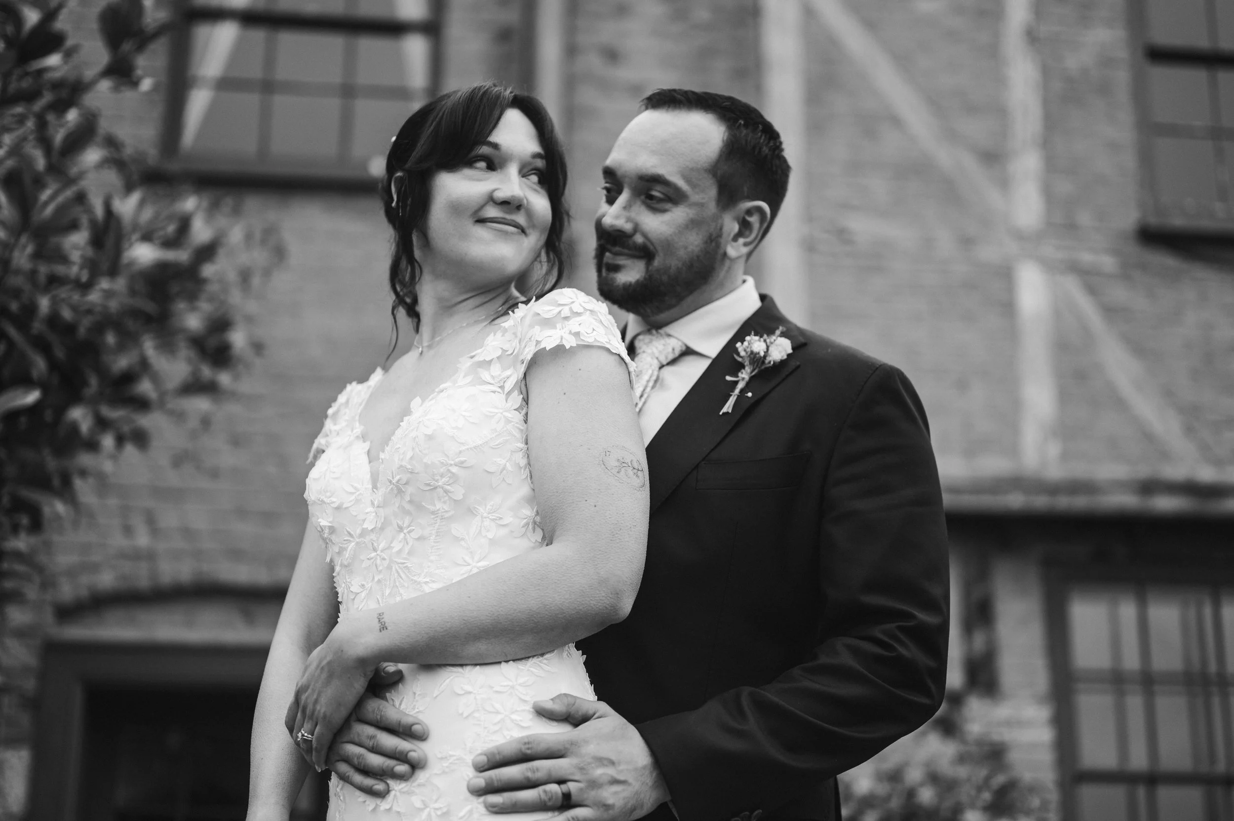 A black and white photo of a smiling bride and groom on their wedding day, standing closely together in front of a brick building, with the groom holding the bride's waist pauntley court, Gareth roy photography