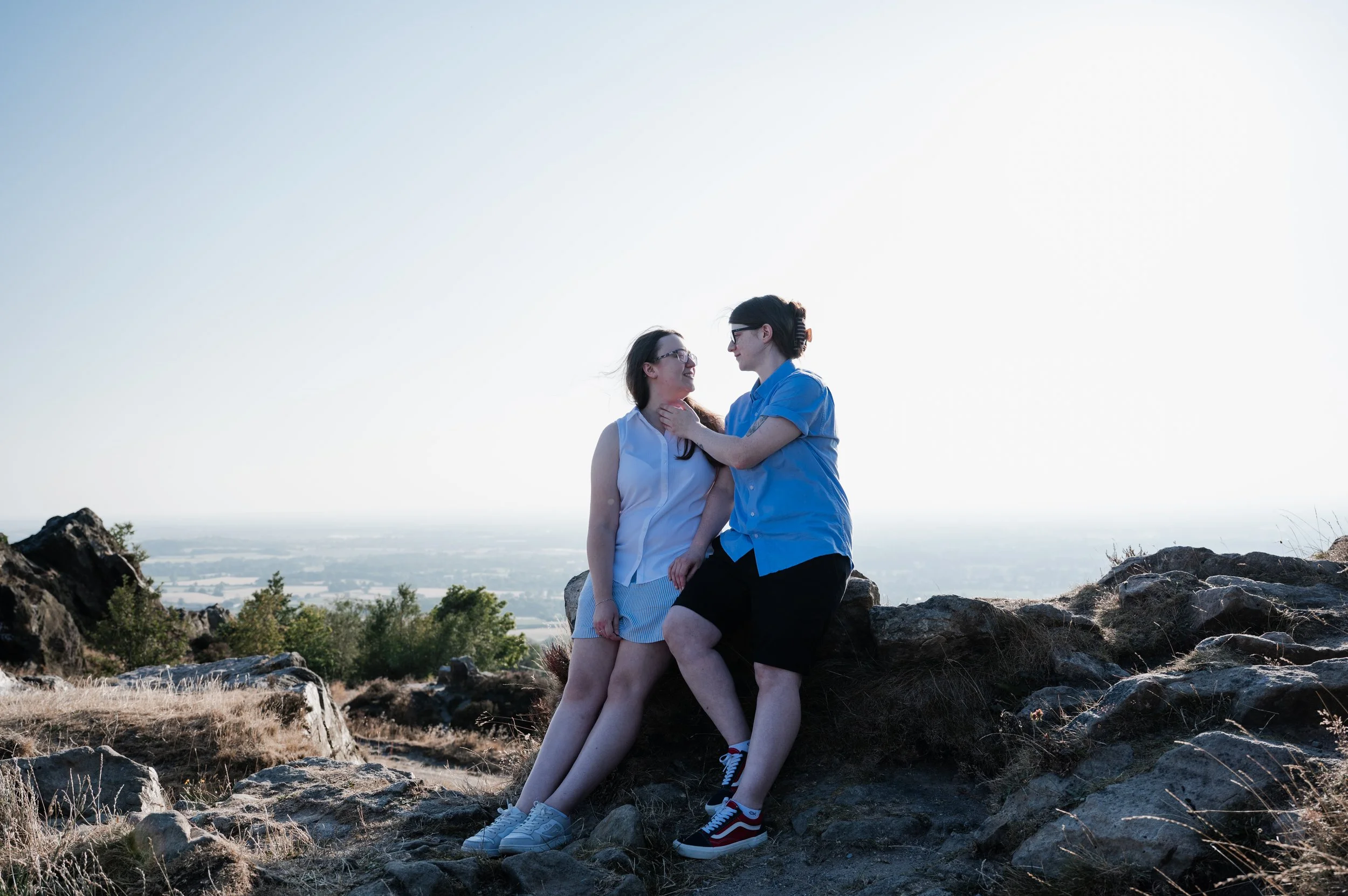 Mow Cop Engagement Shoot - two women stood sitting on a rock with view of Cheshire behind them. 
