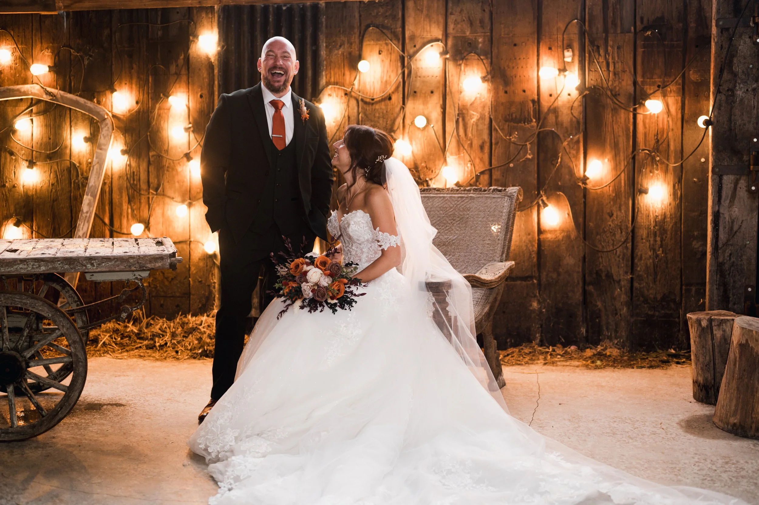 Cheshire Wedding Photographer - Bride sitting in wedding dress and groom standing in a dark suit while laughing at the camera with a background of wooden slats and fairy lights. 