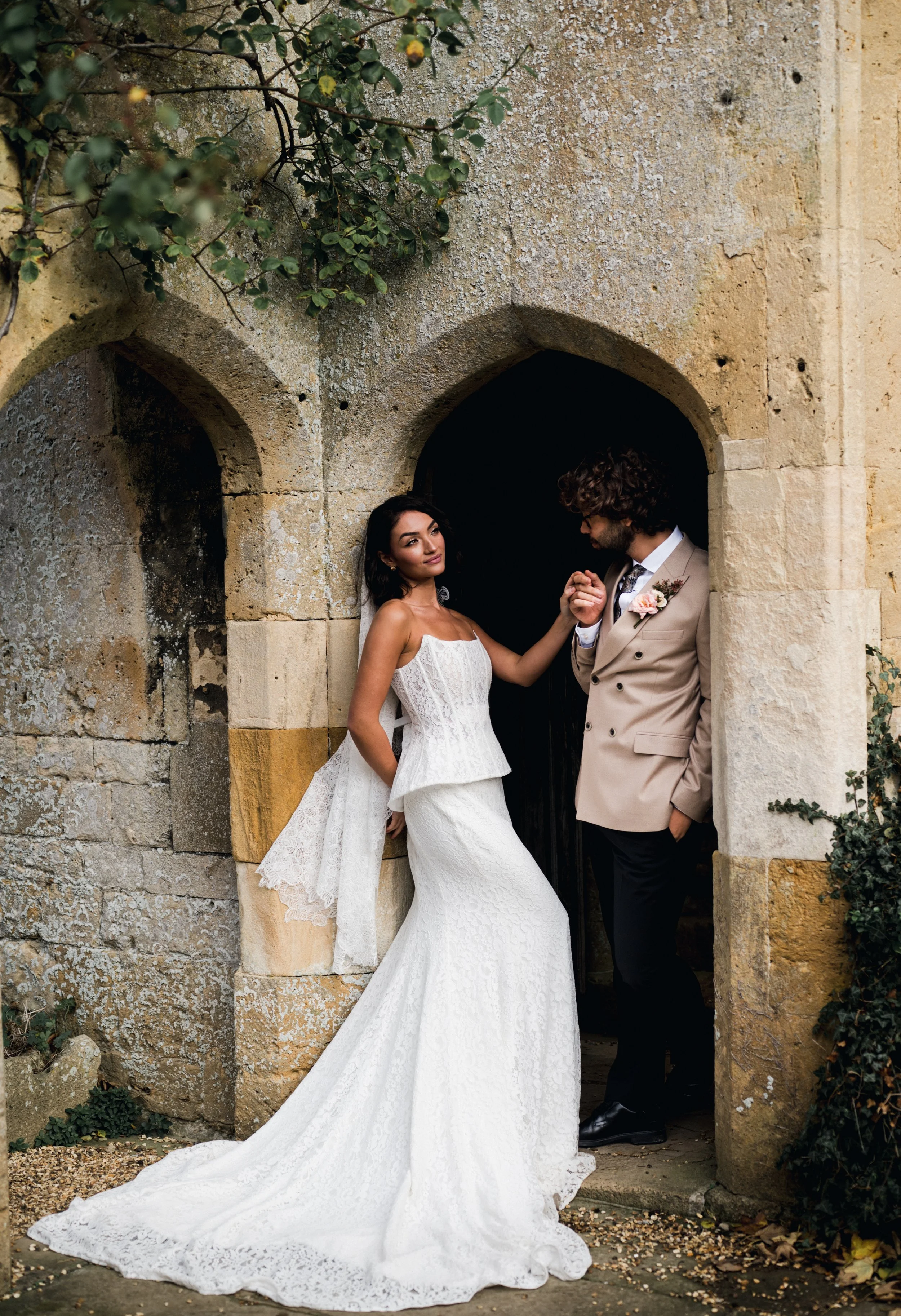 Bride in a white lace wedding dress holding hands with groom in a beige suit beneath an arched stone doorway at sudeley castle, Gareth roy photography