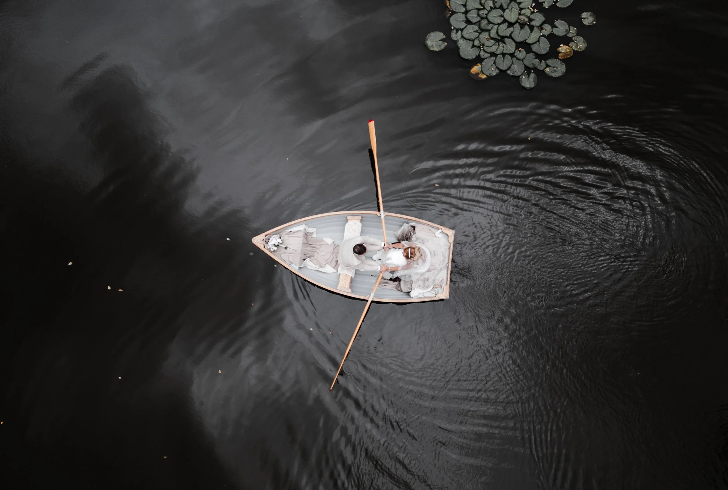 The Love Shack - Drone image of groom and bride in wedding attire, sat in a wooden boat in a lake. 