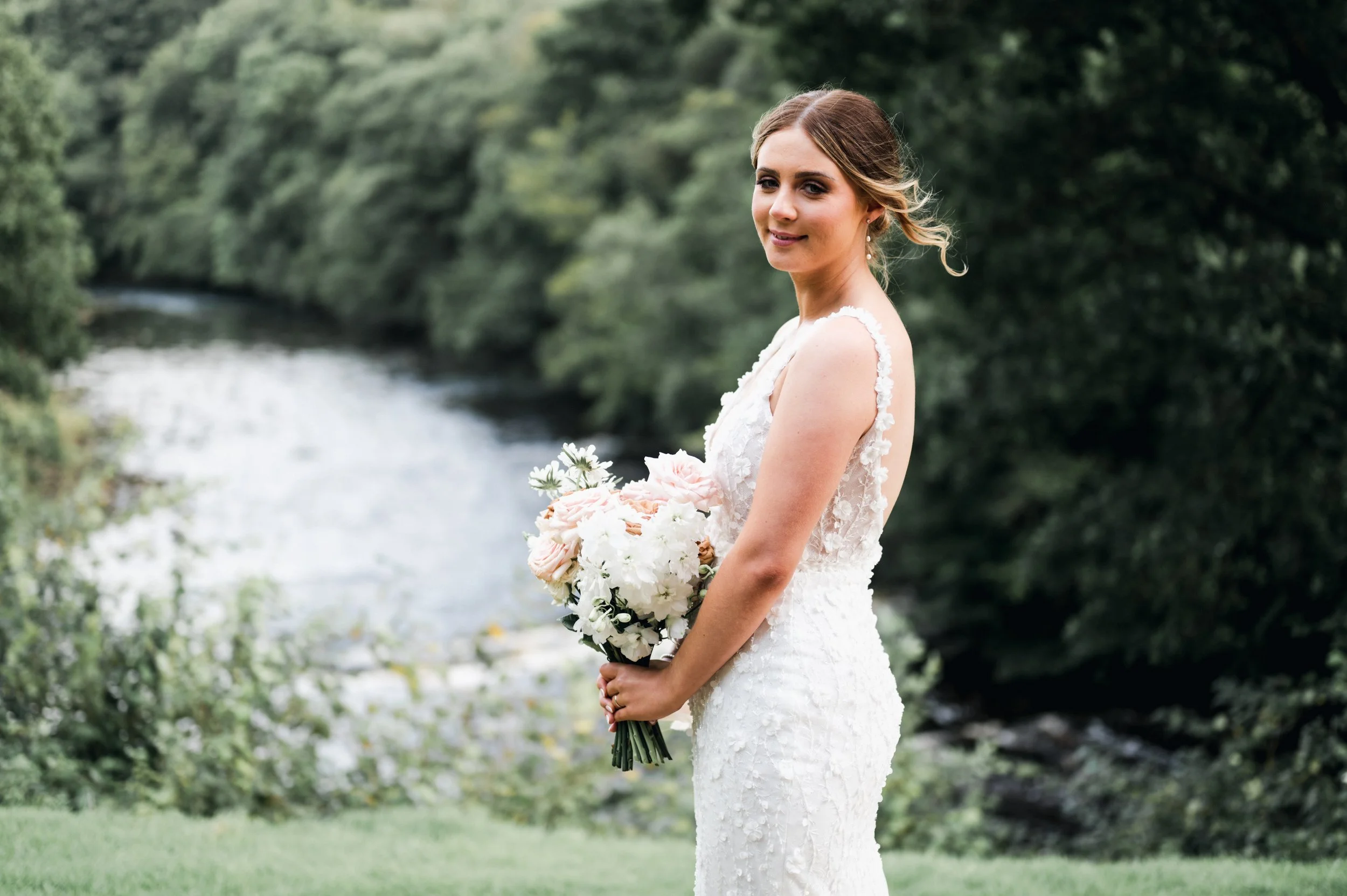 A bride in a white lace wedding dress holding a bouquet of flowers standing outdoors with trees and a river in the background hidden river cabins, Gareth roy photography