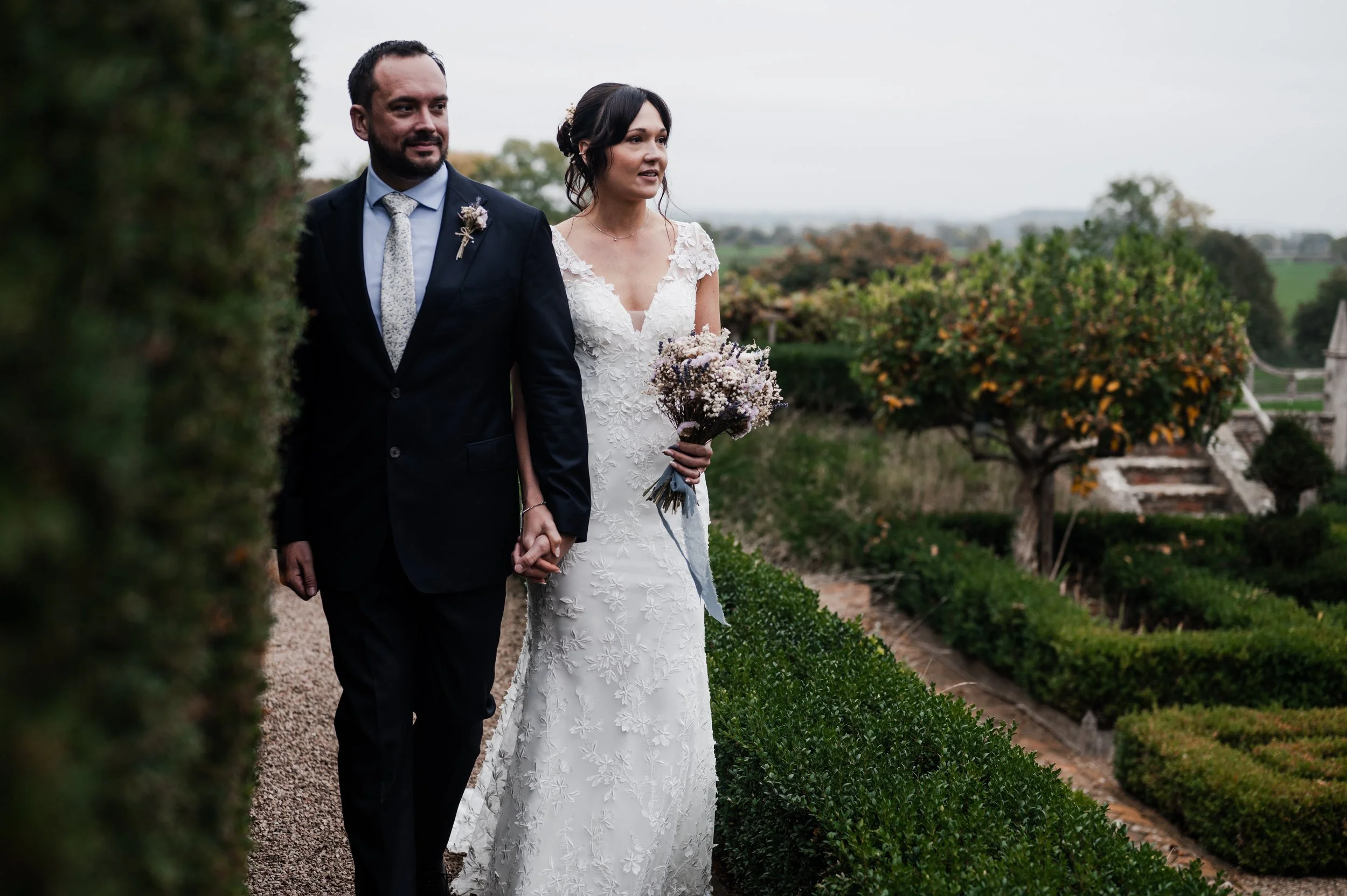 Bride and groom walking hand in hand outdoors in a garden, with the bride holding a bouquet of flowers pauntley court, Gareth roy photography