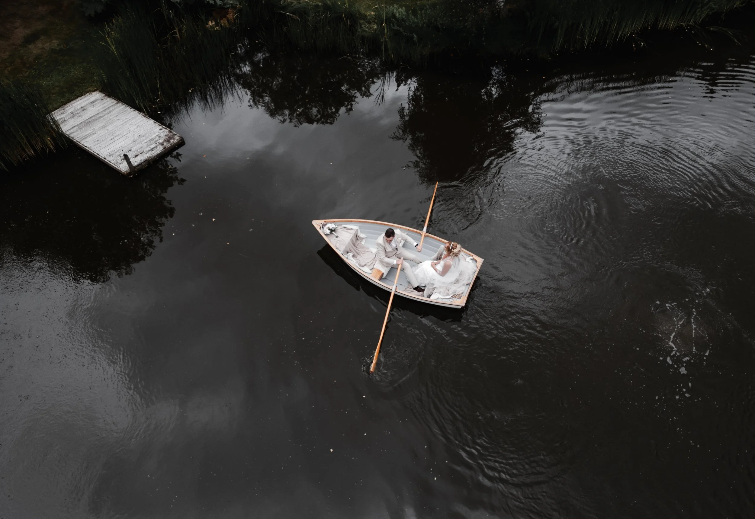 The Love Shack - Drone image of groom and bride in wedding attire, sat in a wooden boat in a lake. 