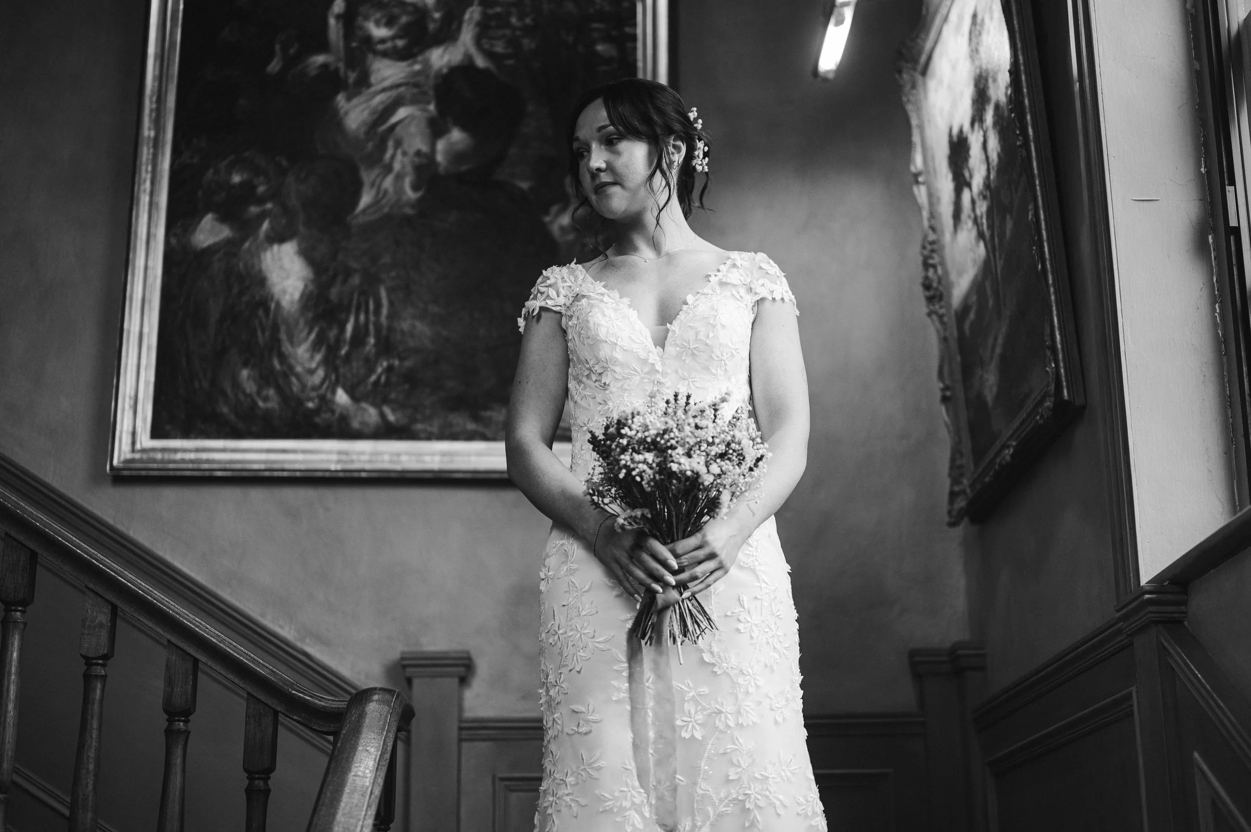 A woman in a wedding dress holding a bouquet standing near a staircase with paintings on the wall pauntley court, Gareth roy photography