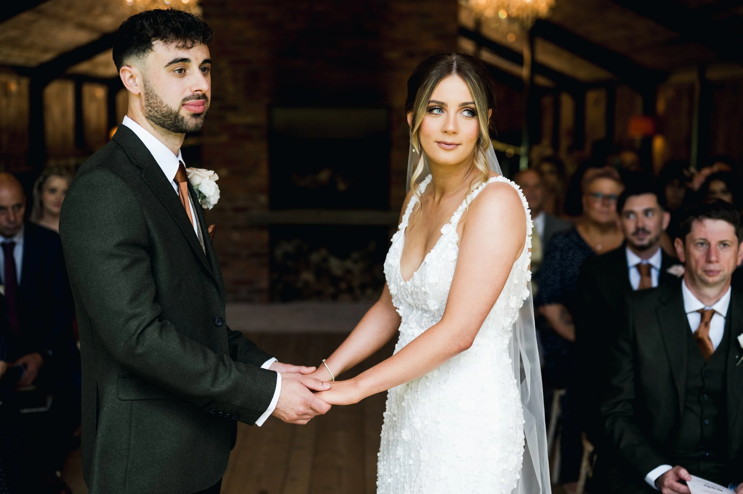 A bride and groom holding hands during their wedding ceremony, surrounded by seated guests in an indoor setting hidden river cabins, Gareth roy photography