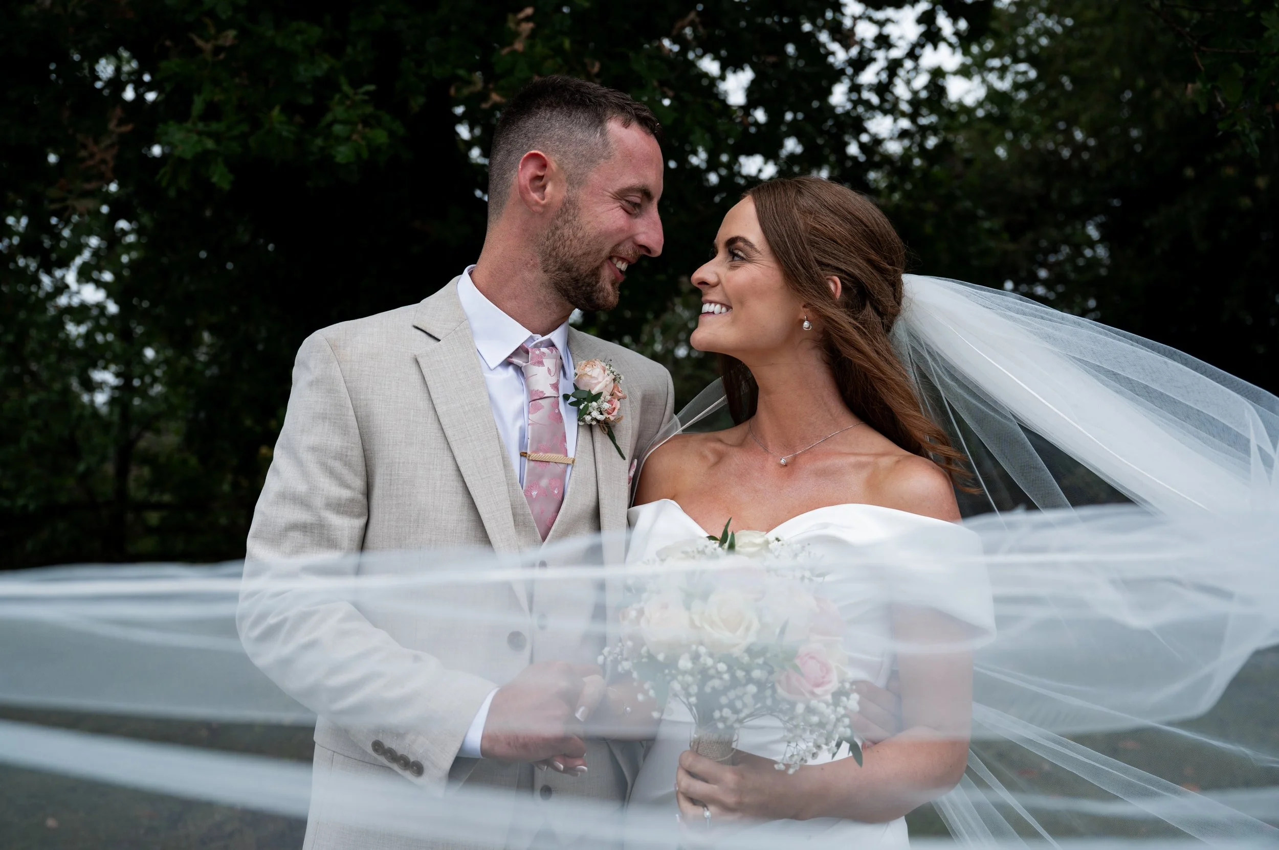 All Saint Church - bride and groom in wedding attire smiling at each other while veil is across the image