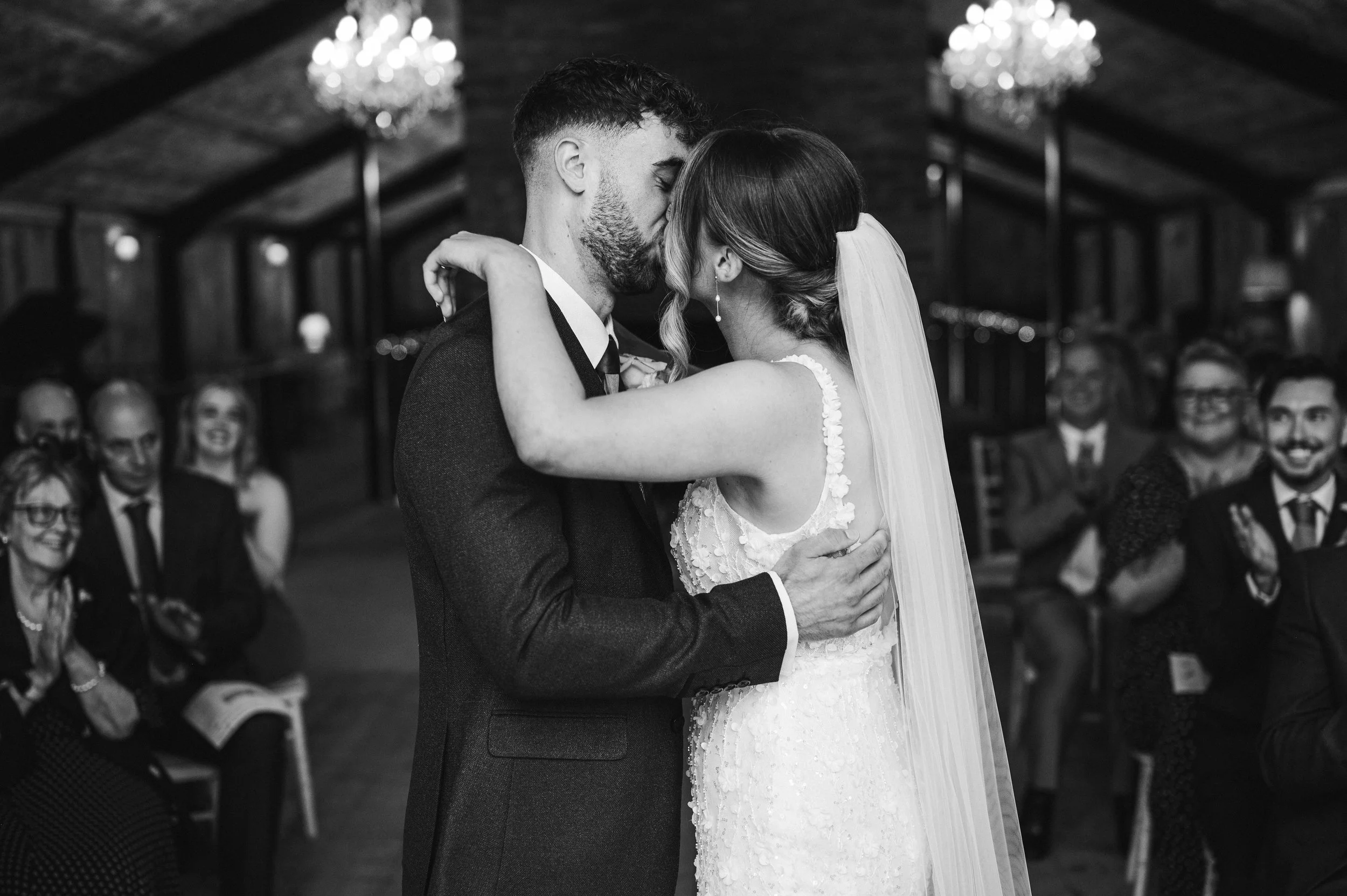 A bride and groom share a kiss during their wedding ceremony in a rustic venue, with guests smiling and clapping in the background hidden river cabins, Gareth roy photography